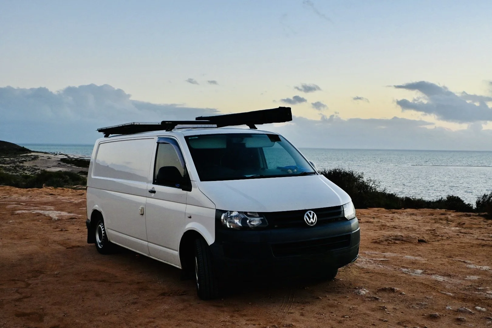 White Volkswagen camper van parked on a dirt patch near the ocean at sunset, with a surfboard secured on top.