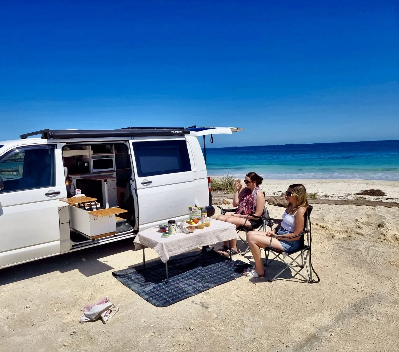 Two women sit in camping chairs on a sandy beach, having a picnic near a white camper van with an open door and a small table with food and drinks. The ocean and blue sky are in the background.