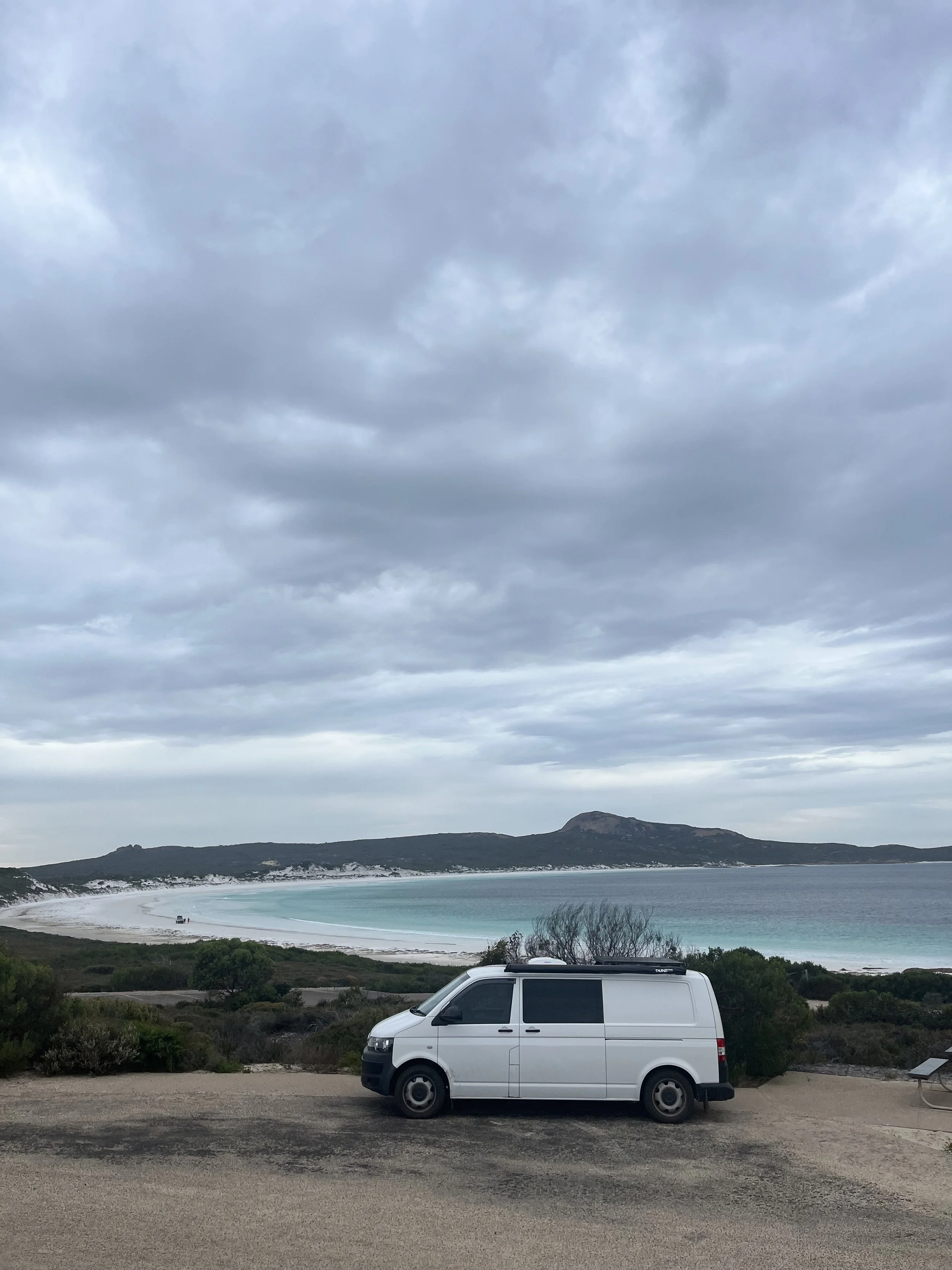 A white van parked on a dirt road near a beach with a cloudy sky and a mountain in the background.