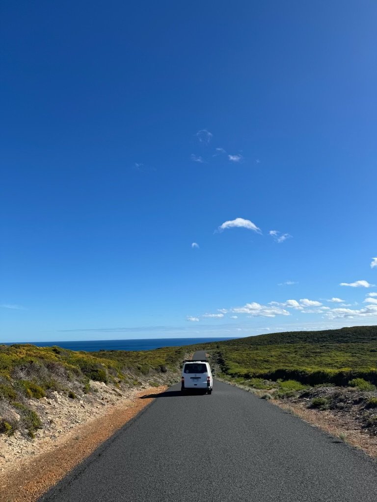 A white van driving down a straight, empty road through open landscape under a bright blue sky with scattered clouds, ocean visible in the distance.
