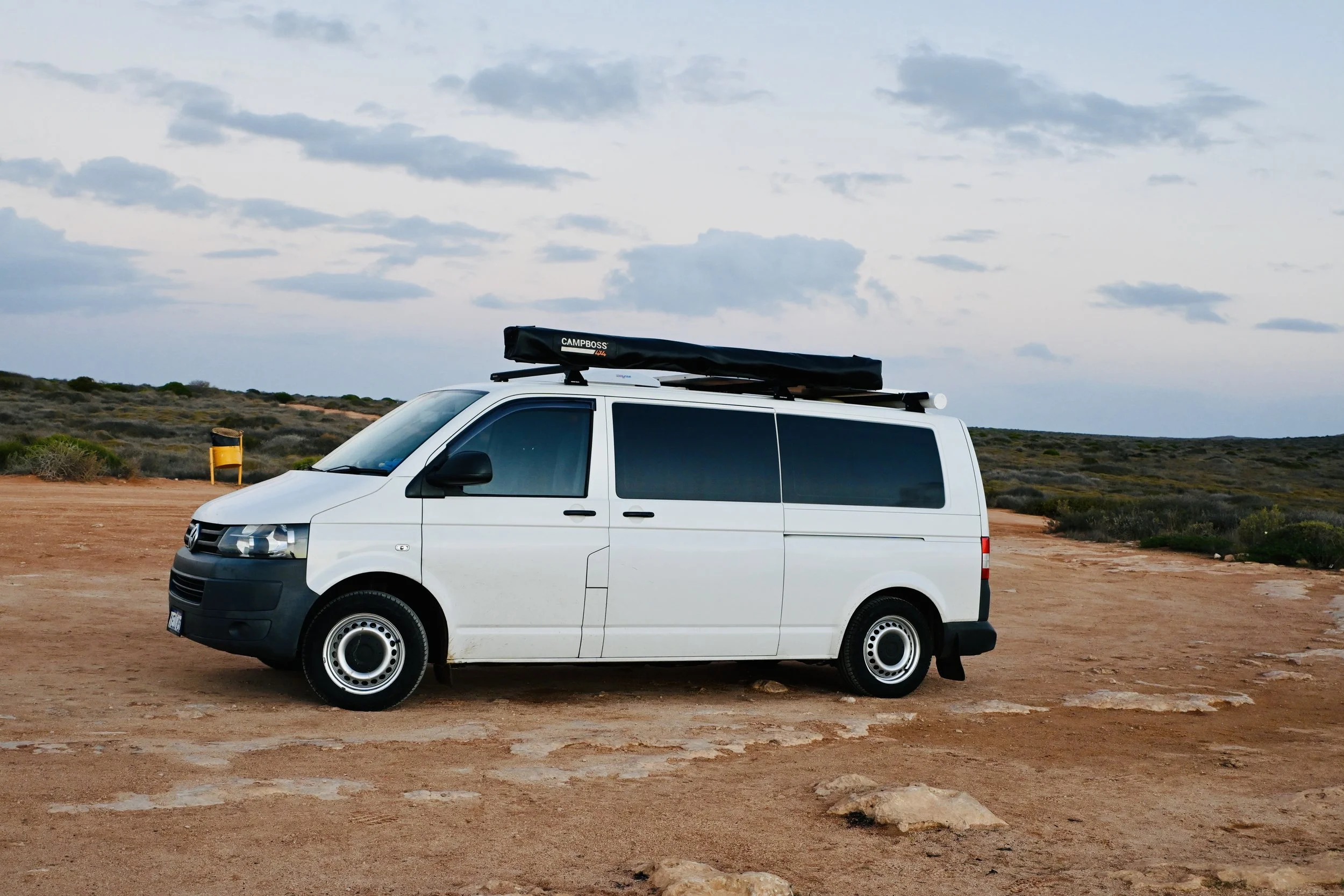 White camper van parked on a dirt area in a semi-arid landscape with low vegetation under a cloudy sky.