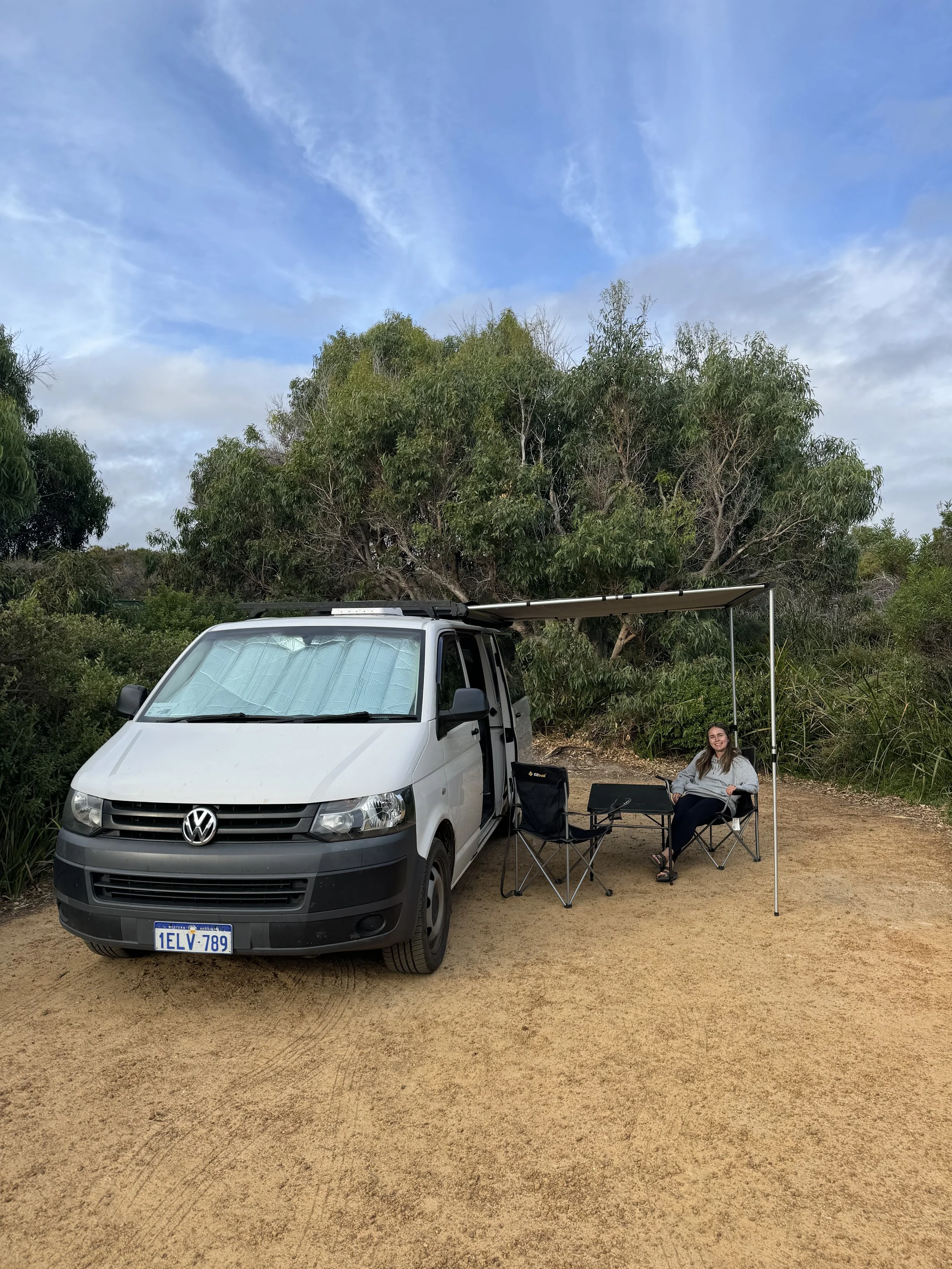 A woman sitting on a camping chair beside a white Volkswagen van with a sunshade, on a dirt patch surrounded by trees and bushes, under a blue sky with some clouds.
