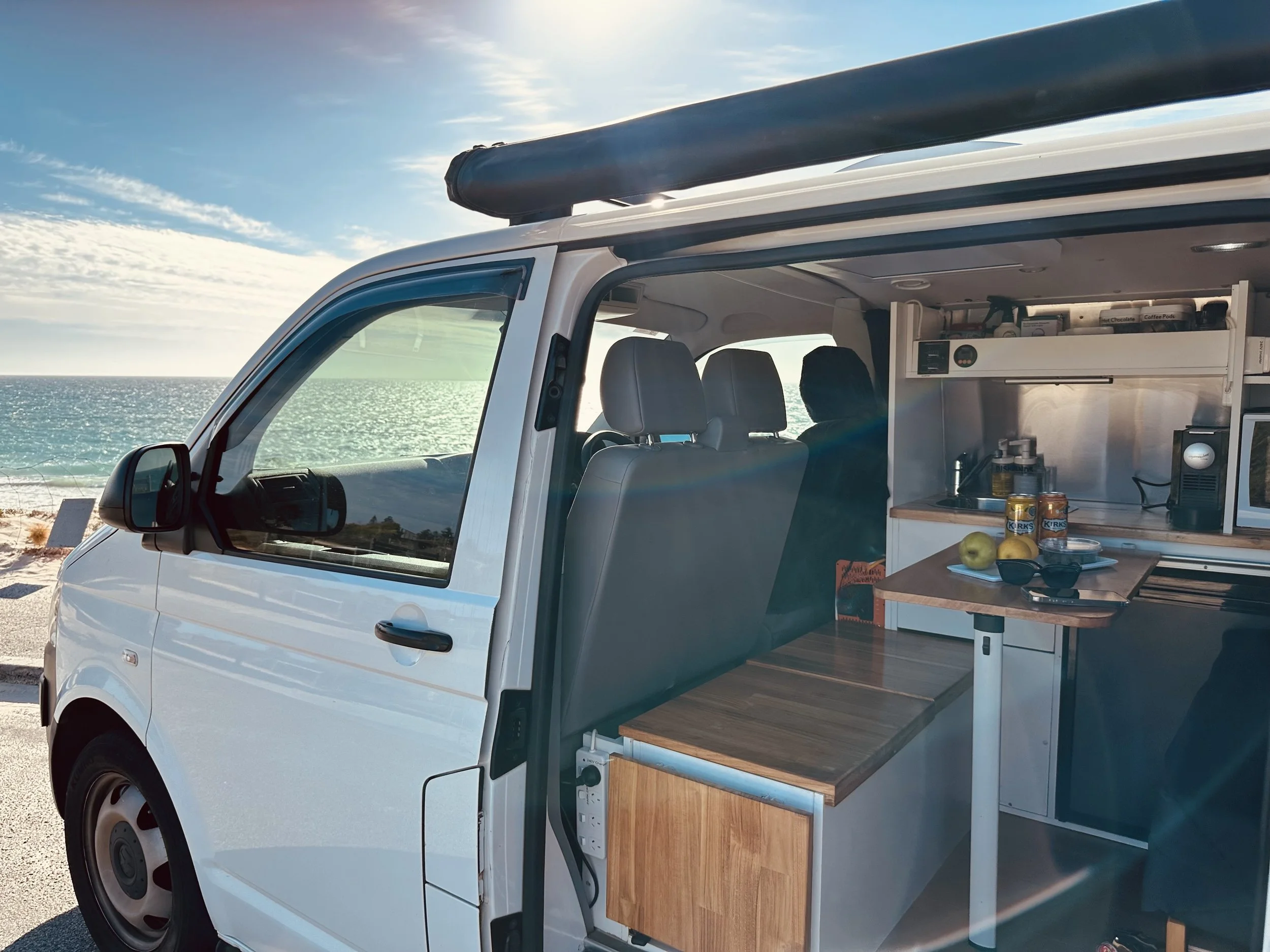 A white camper van parked by the beach with the door open, showing the interior kitchen with apples, canned goods, and kitchen appliances, overlooking the ocean under a partly cloudy sky.