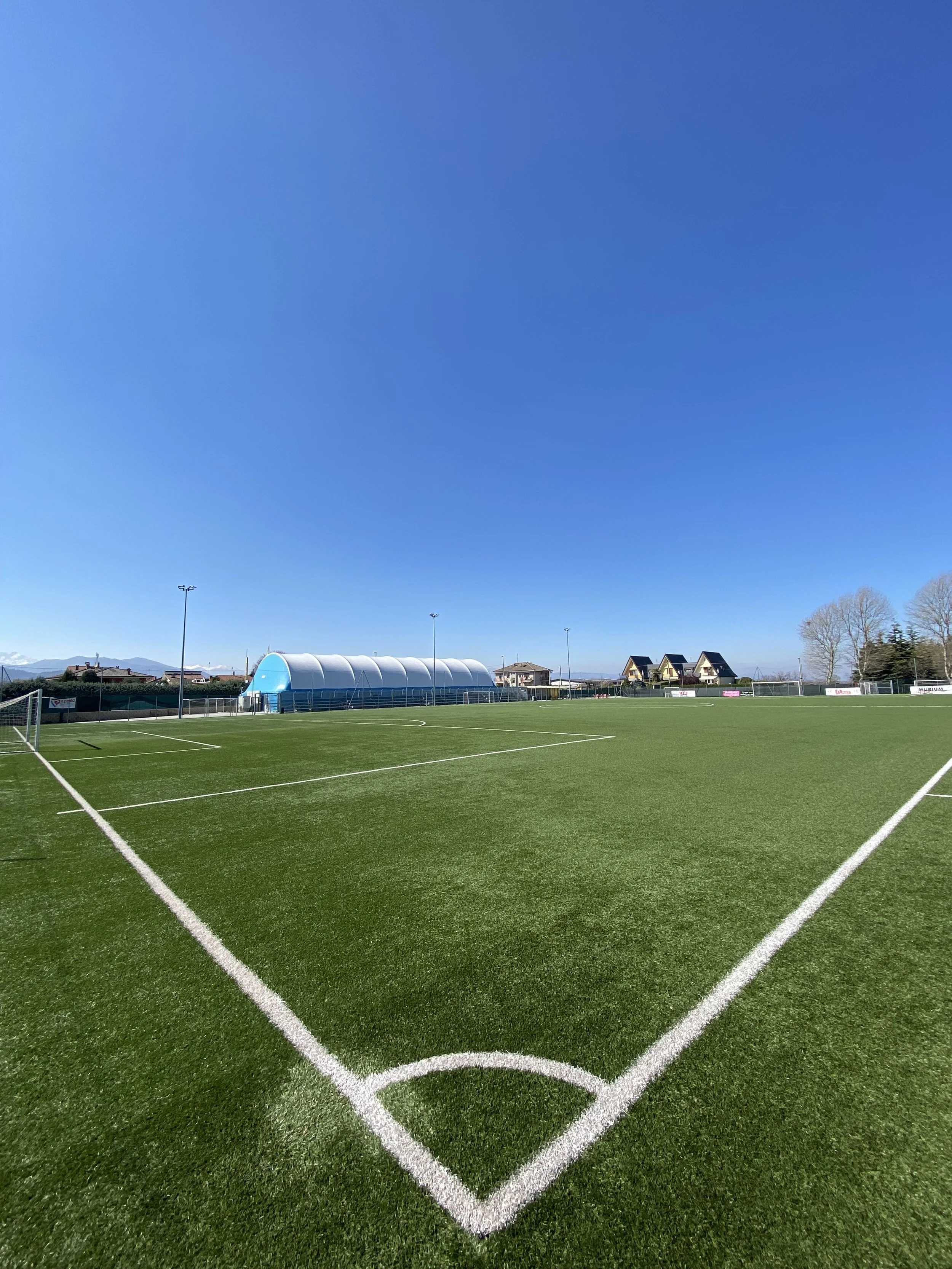 Campo di calcio con erba verde e linee bianche, cielo blu, edifici in lontananza.