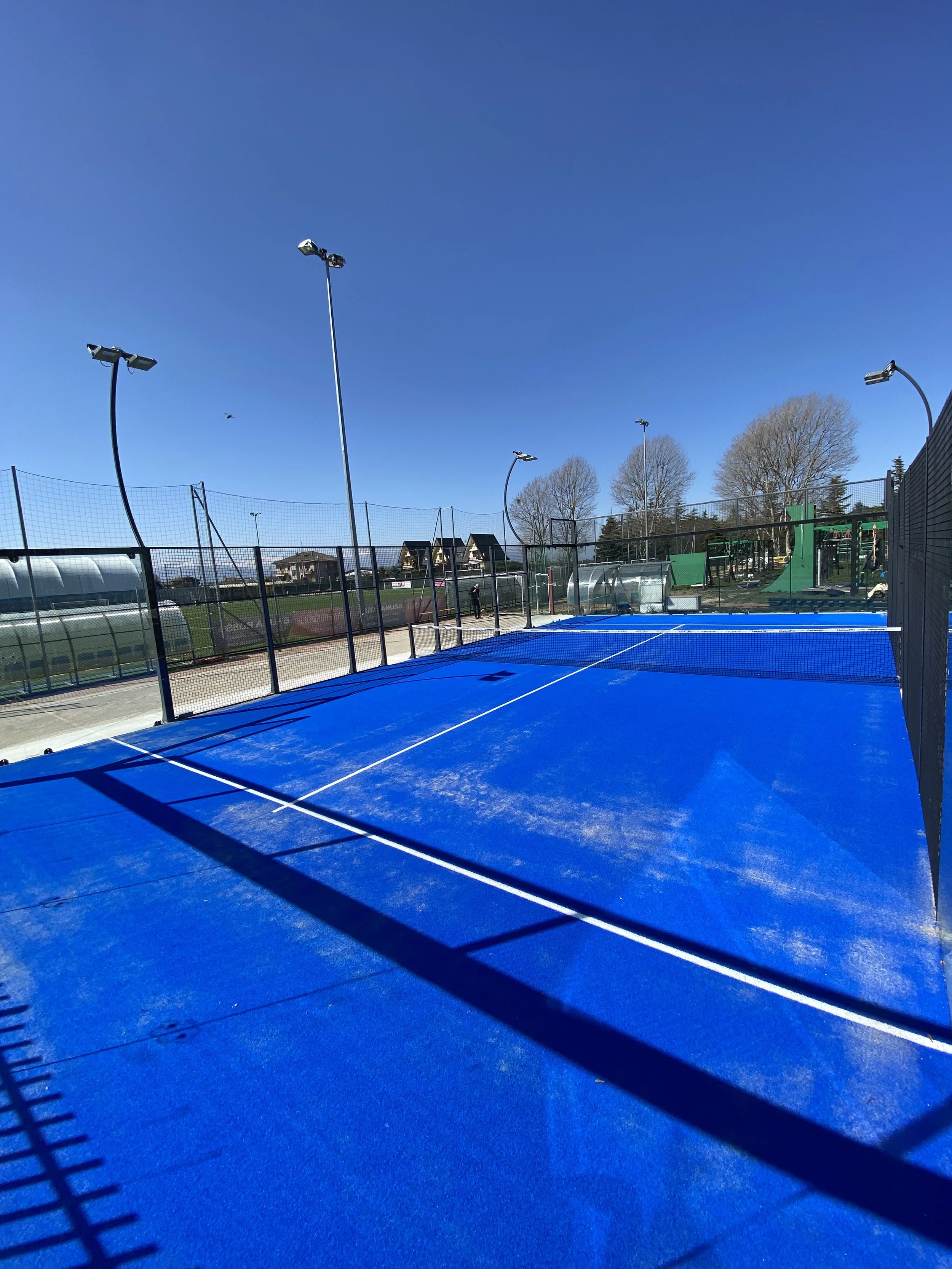 Campo da padel all'aperto con erba blu e rete, racchiuso da recinzioni e con aree verdi e alberi sullo sfondo.