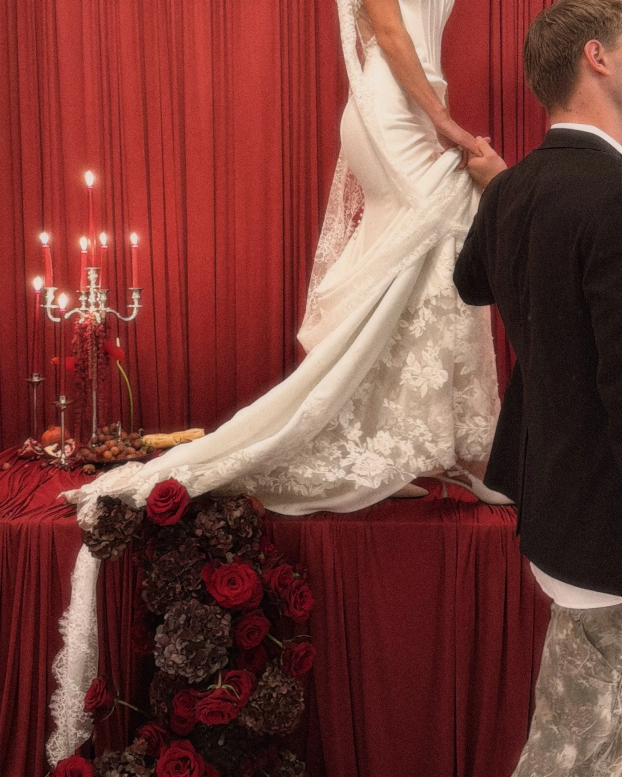 Moody wedding aesthetic featuring a bride with a long lace train standing on a table with red roses and candles. High-fashion, behind-the-scenes content captured for social media