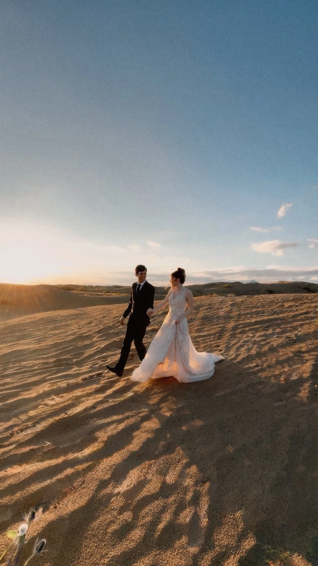 A bride and groom walking hand in hand across sandy desert dunes at sunset, with a bright sky and distant hills.