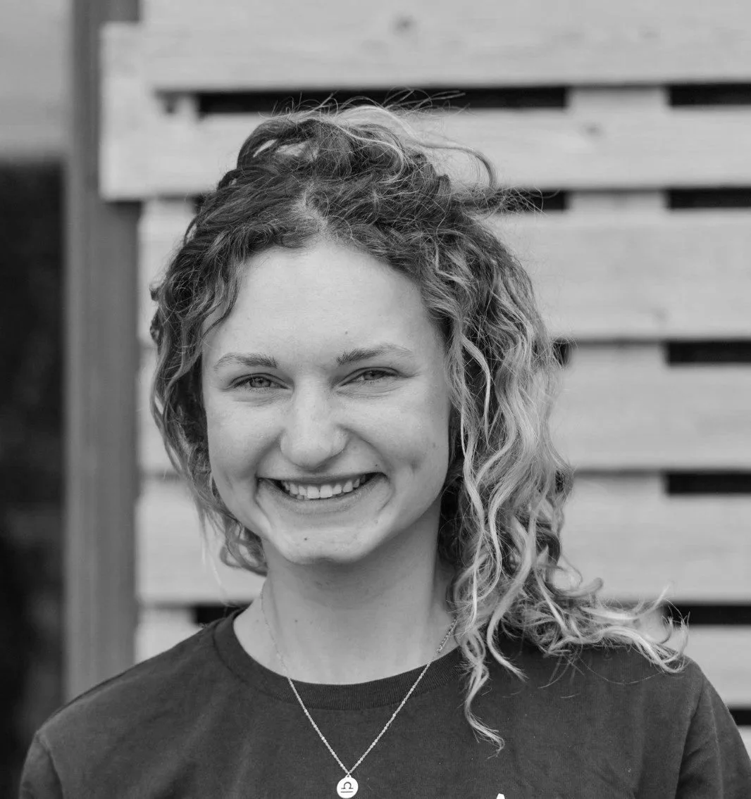 A smiling woman with curly hair wearing a necklace, standing outdoors in front of a wooden fence.