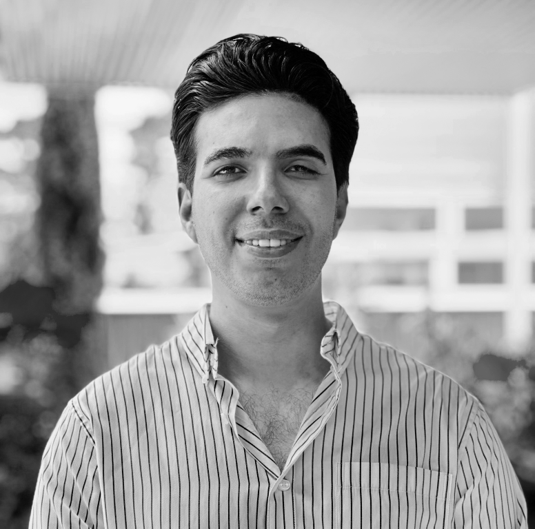 Young man with dark hair wearing a striped button-down shirt, smiling in an outdoor setting with blurred buildings and trees in the background.