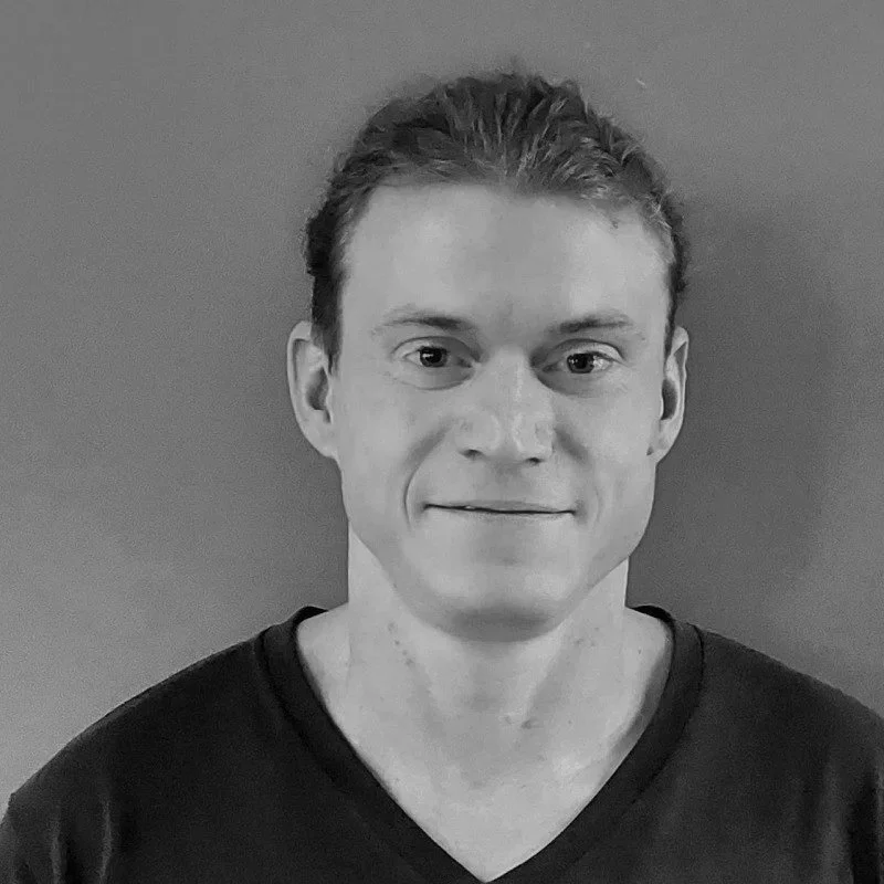 Black and white photo of a young man with dark hair, smiling, wearing a collared shirt, with an indoor background.