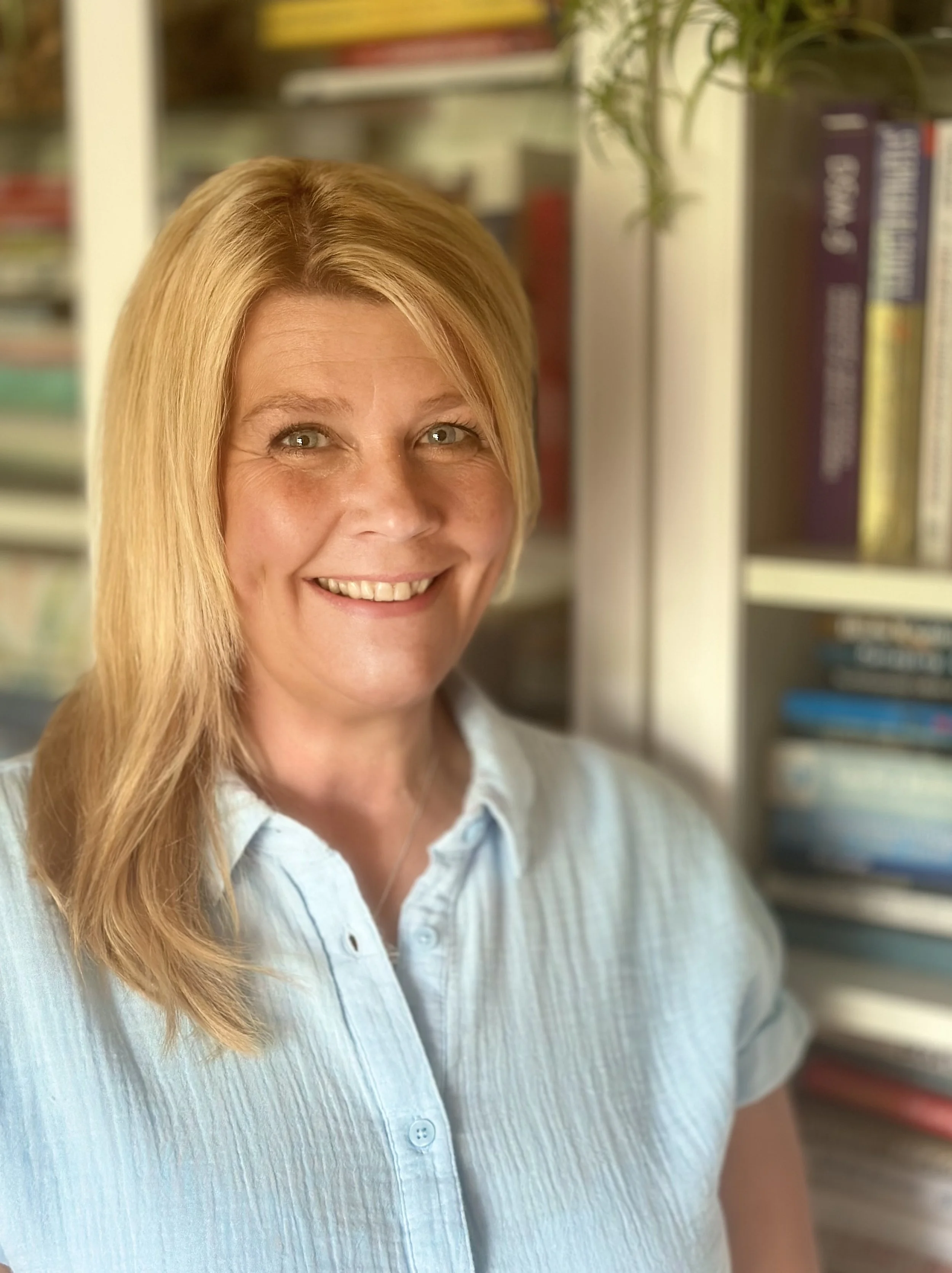 Smiling person with blonde hair in blue shirt standing in front of a bookshelf.