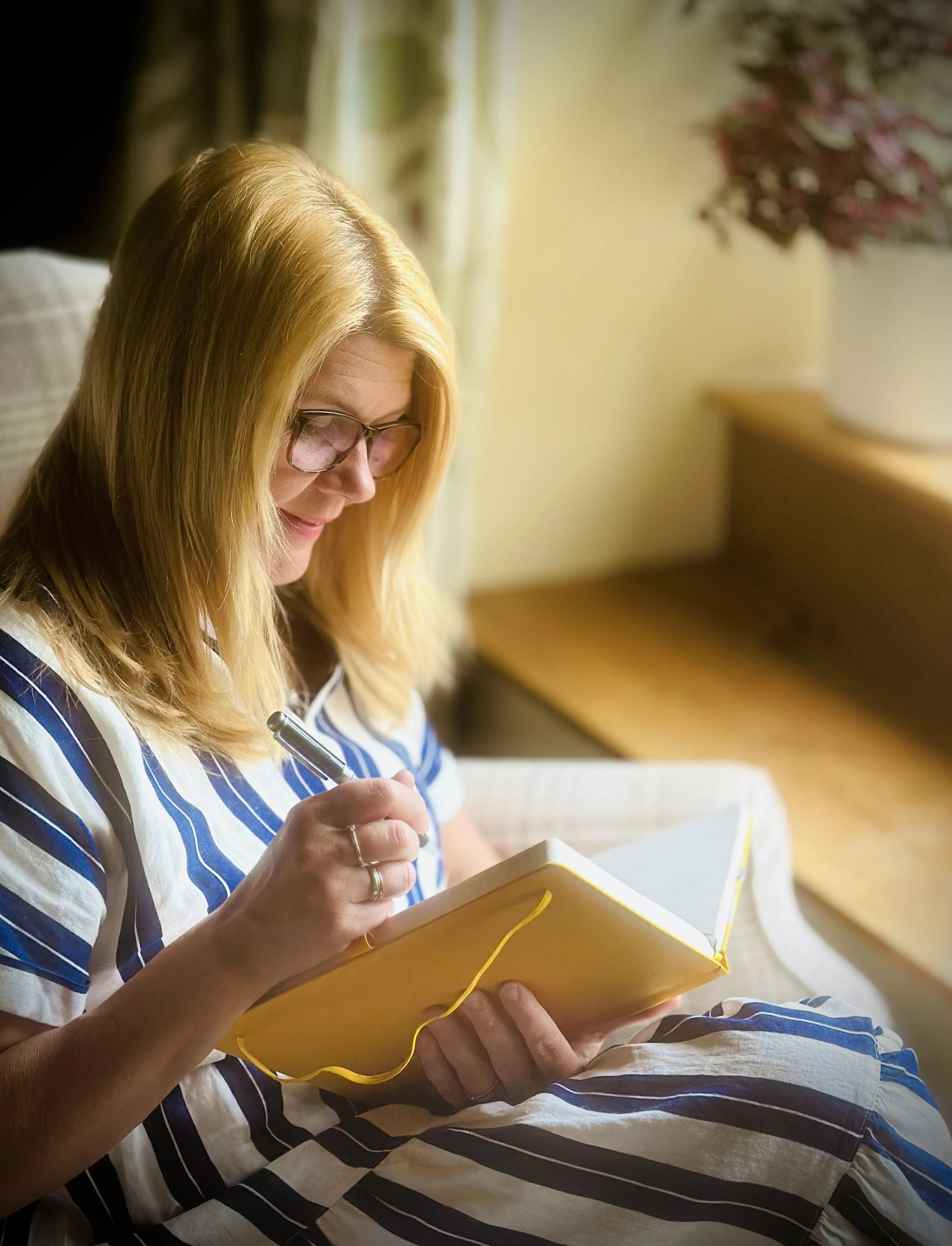 Woman with blonde hair wearing glasses, sitting indoors in a sunlit room, writing in a yellow notebook.