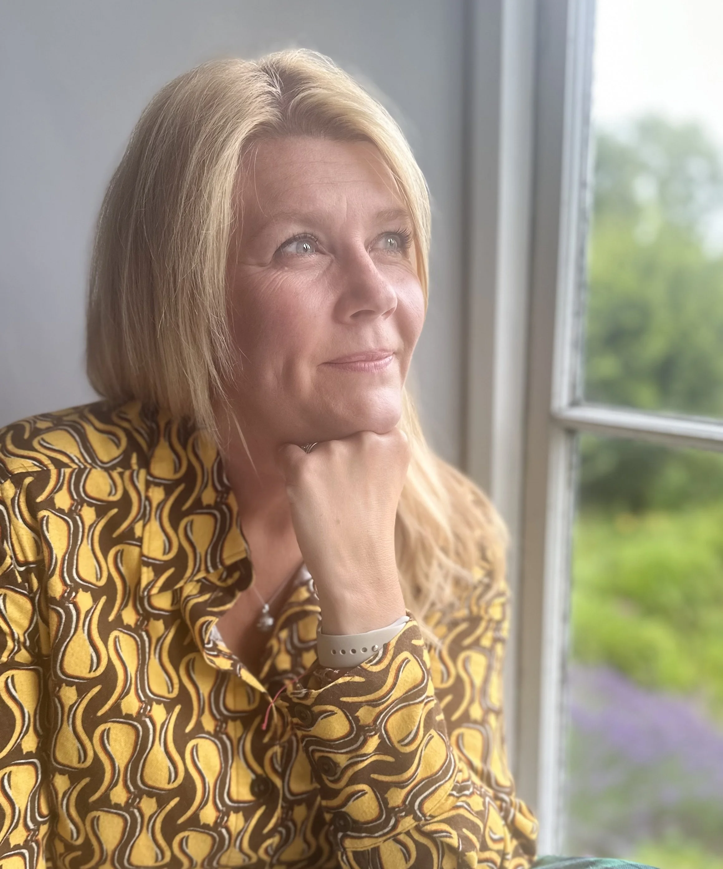 A woman with blonde hair, wearing a patterned yellow shirt, is looking thoughtfully out of a window, with her chin resting on her hand.