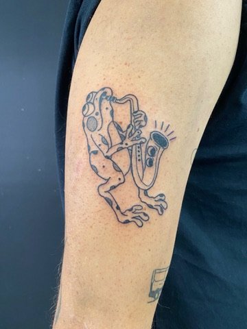 Tattoo of a frog playing a saxophone on a person's upper arm.