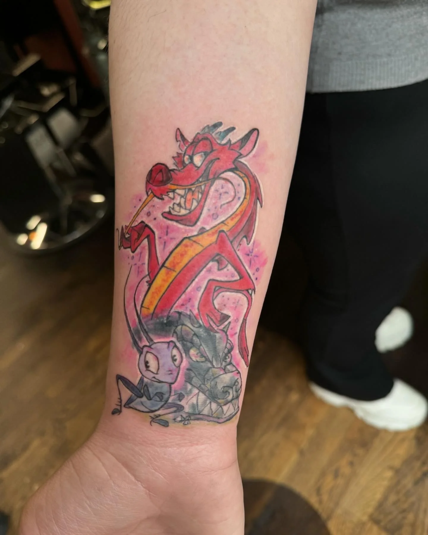 Colorful tattoo of Disney characters on person's forearm, featuring Mushu, Mulan's dragon, and small characters Mushu, Cri-Kee, and Dragon from Disney animated film.