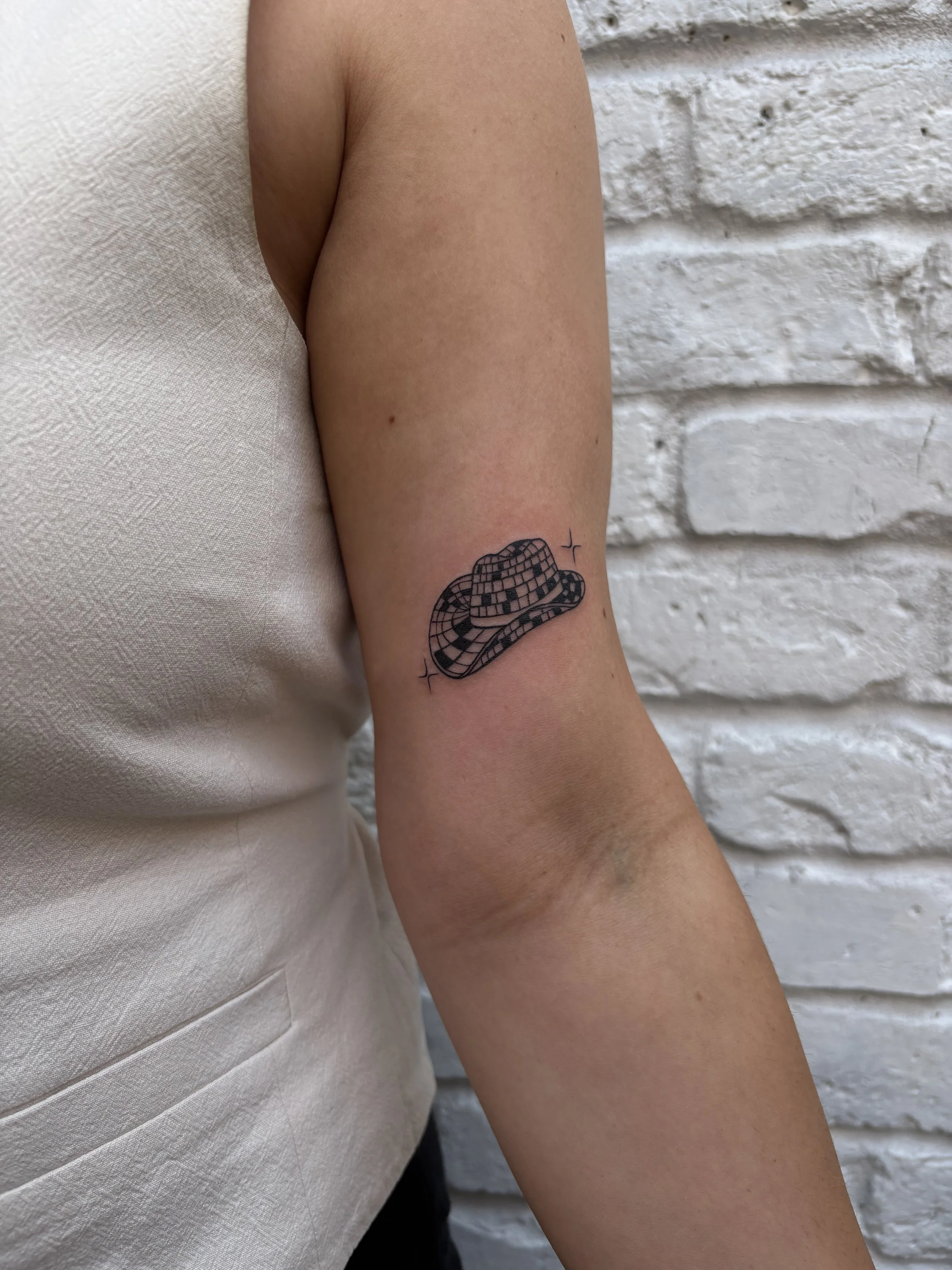 A person's upper arm with a tattoo of a checkered cowboy hat on the inner side, with sparkles around it and a white textured brick wall in the background.