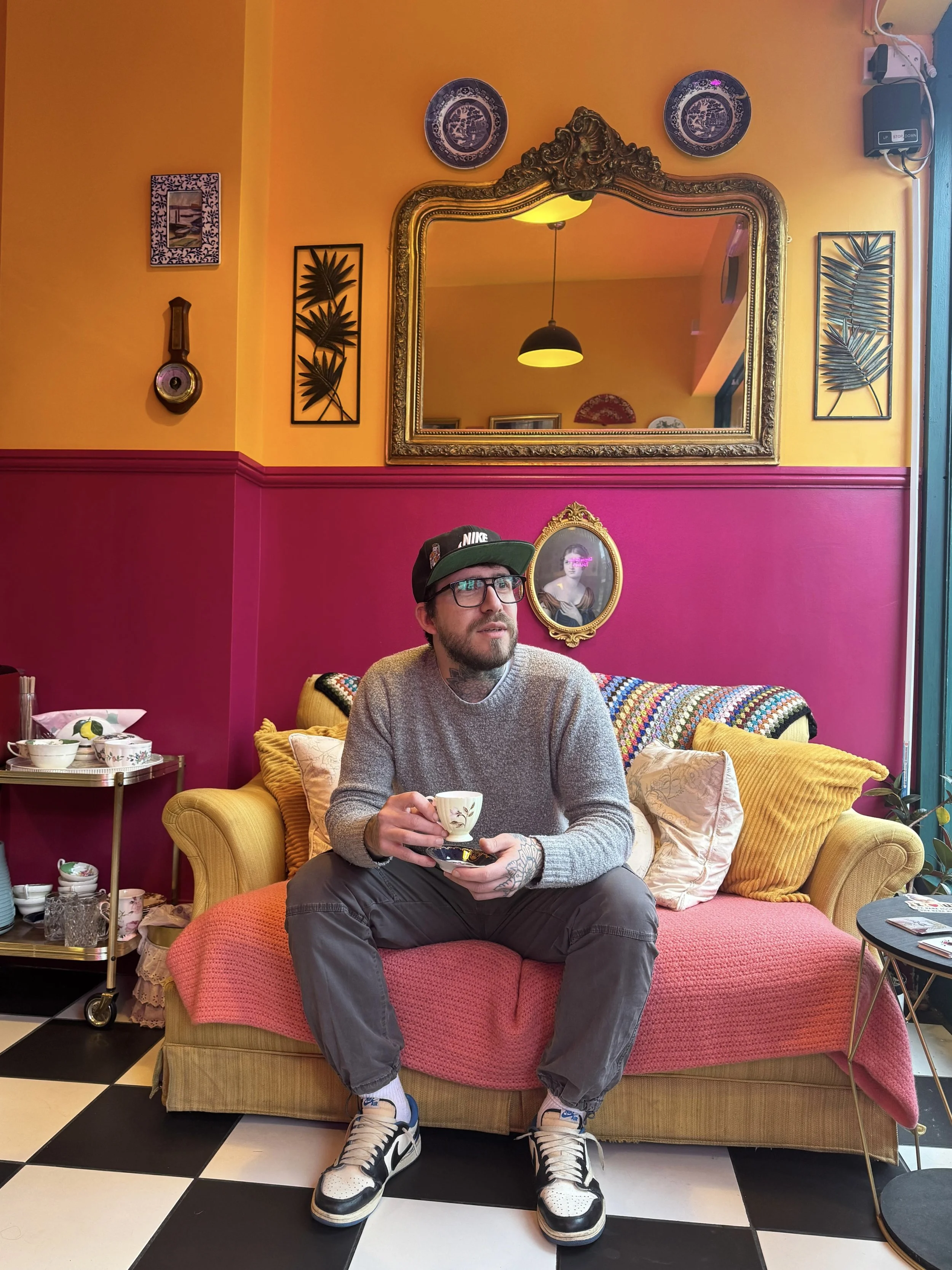 A man with glasses and a beard, wearing a black Nike cap, gray sweater, and sneakers, sitting on a colorful sofa in a vibrant room, holding a cup and saucer, with a wall decorated with ornate mirror, plates, and framed art behind him.