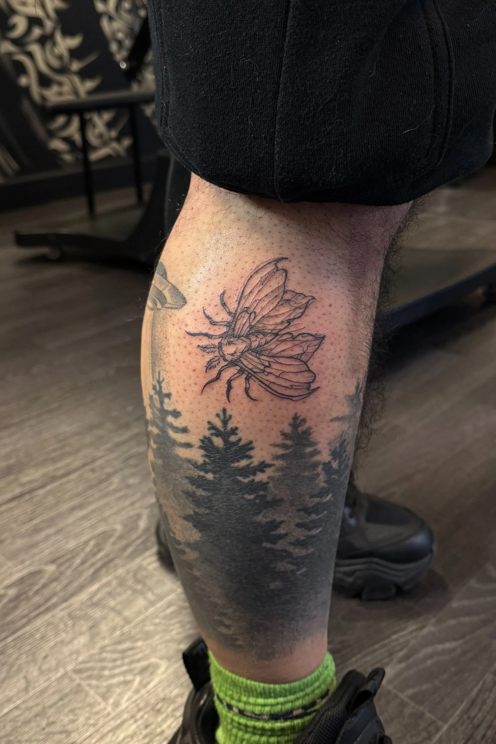 Tattoo of a bee on a person's lower leg, with pine trees depicted below.