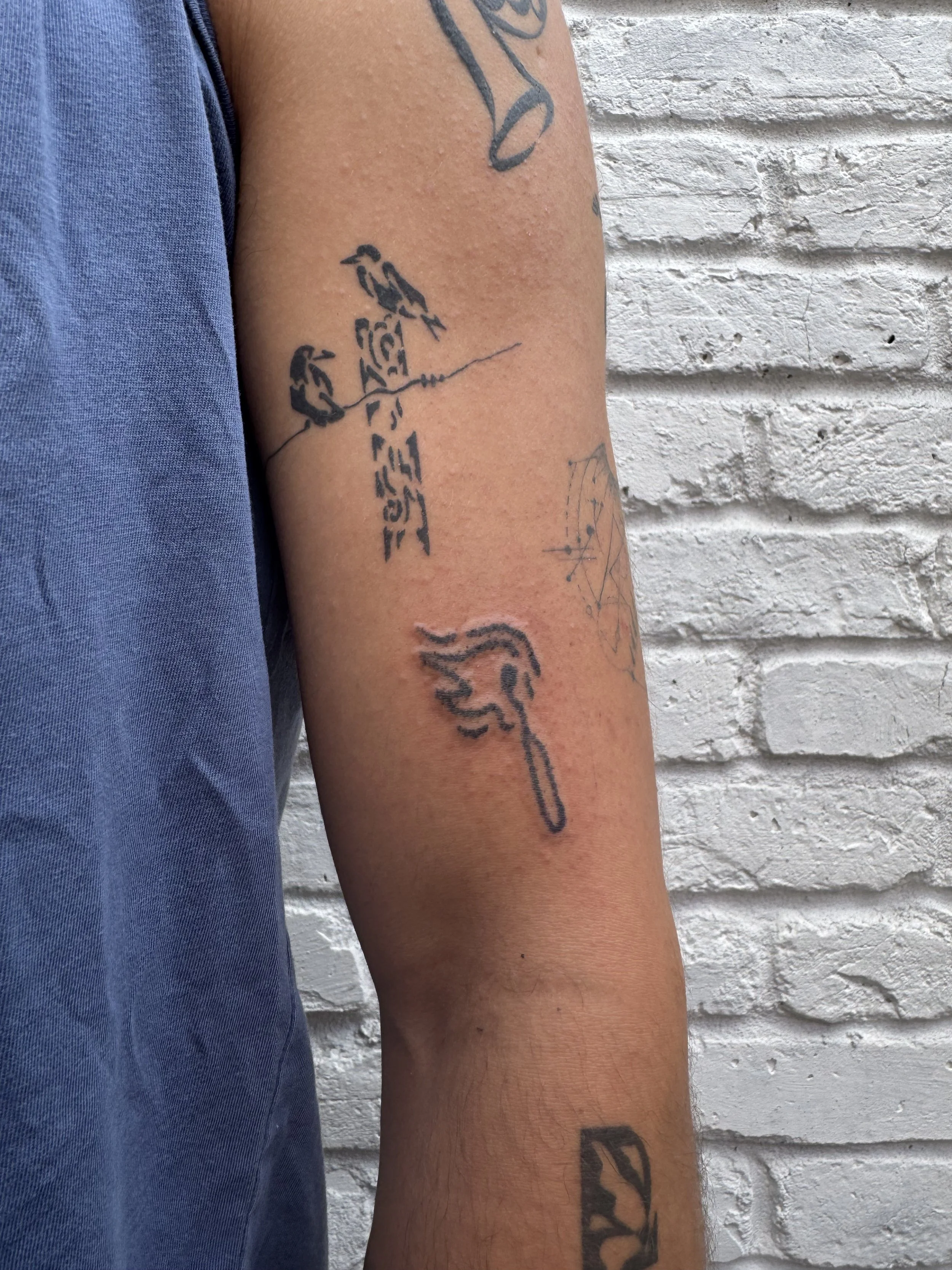 Close-up of a person's arm with several tattoos, including a flame tattoo, a bird tattoo, and a tattoo with text, against a white brick wall background.