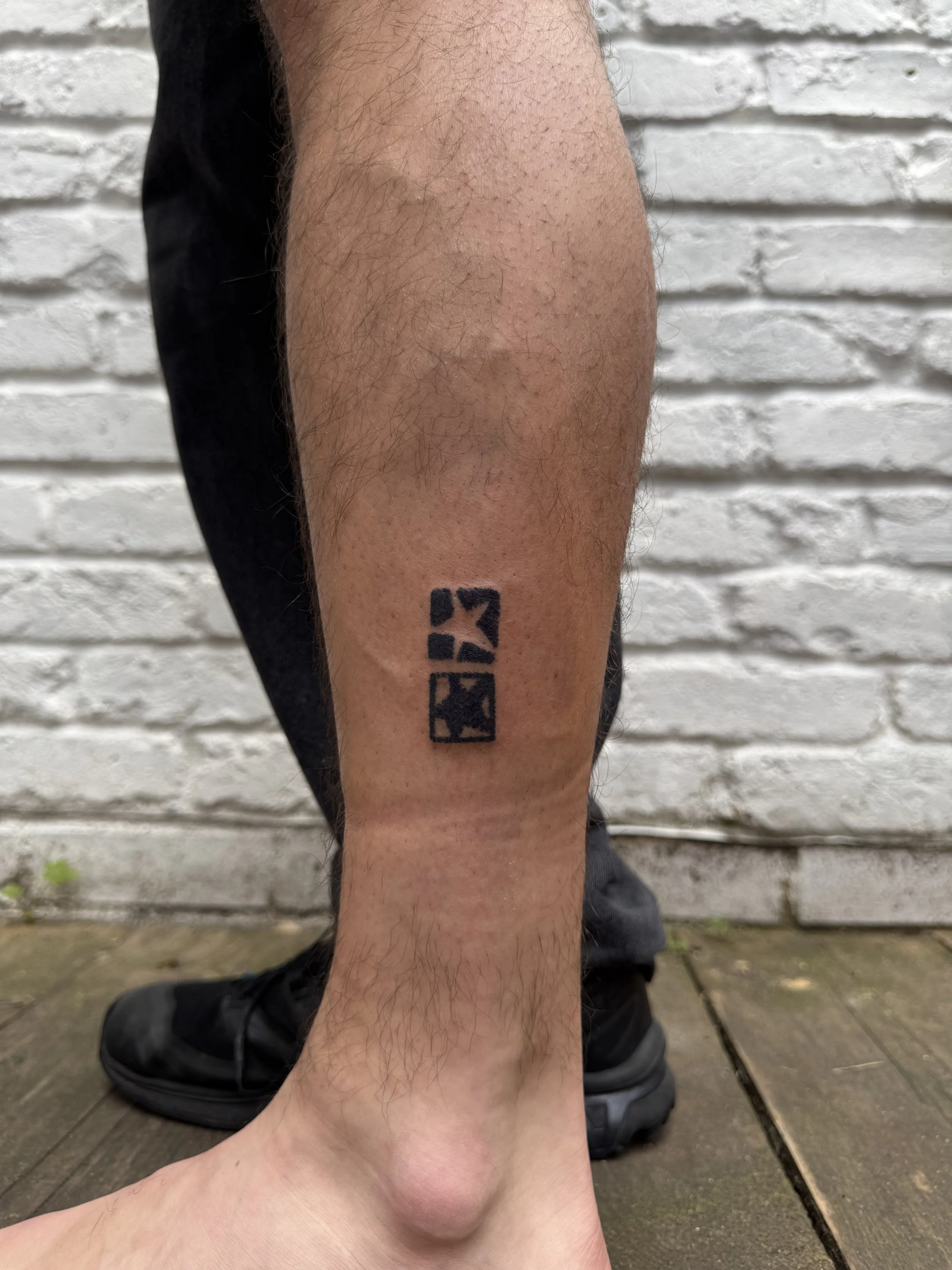 A person's lower leg with a black tattoo of three stylized squares arranged vertically against a white brick wall background.