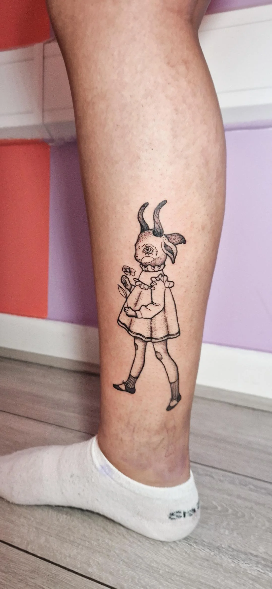 Tattoo of a character with a goat head, a single eye, and human body, holding a flower, on a person's leg.
