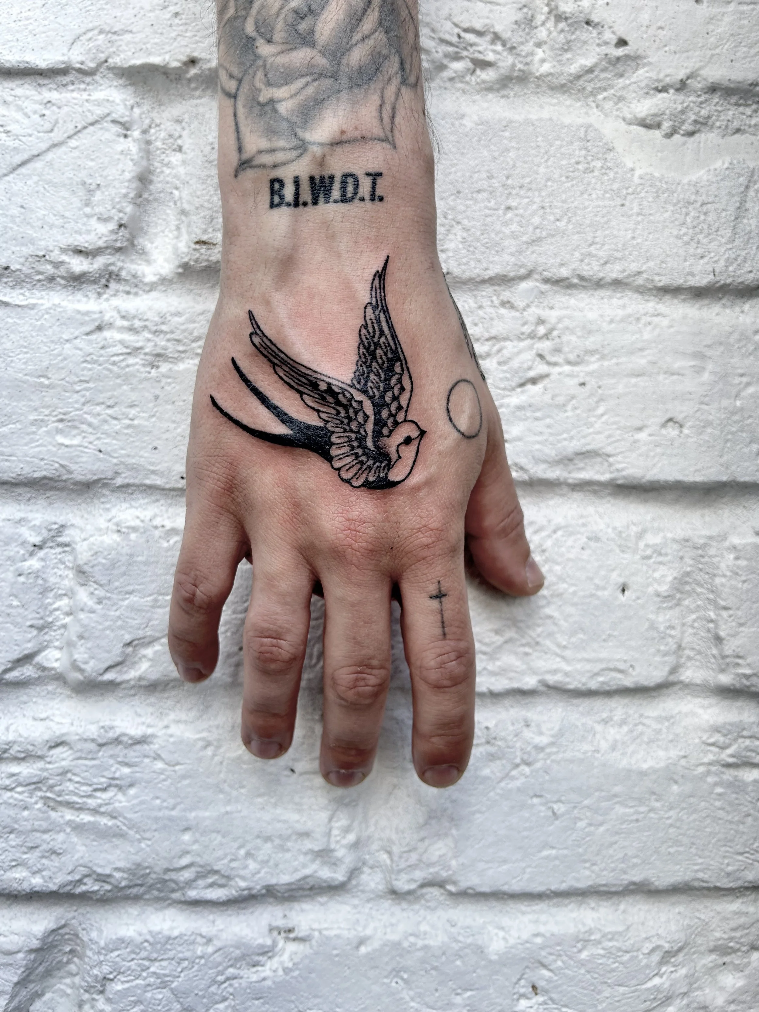 A person’s hand with tattoos including a swallow bird, a sun, a cross on the finger, and the words "B.I.W.D.T." on the wrist, against a white brick wall background.