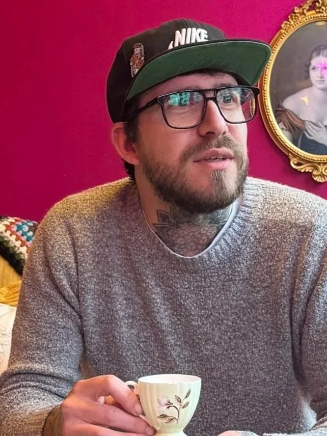 Man with glasses, beard, and tattoos wearing a black Nike cap, holding a teacup, in a room with a pink wall and a framed portrait of a woman.