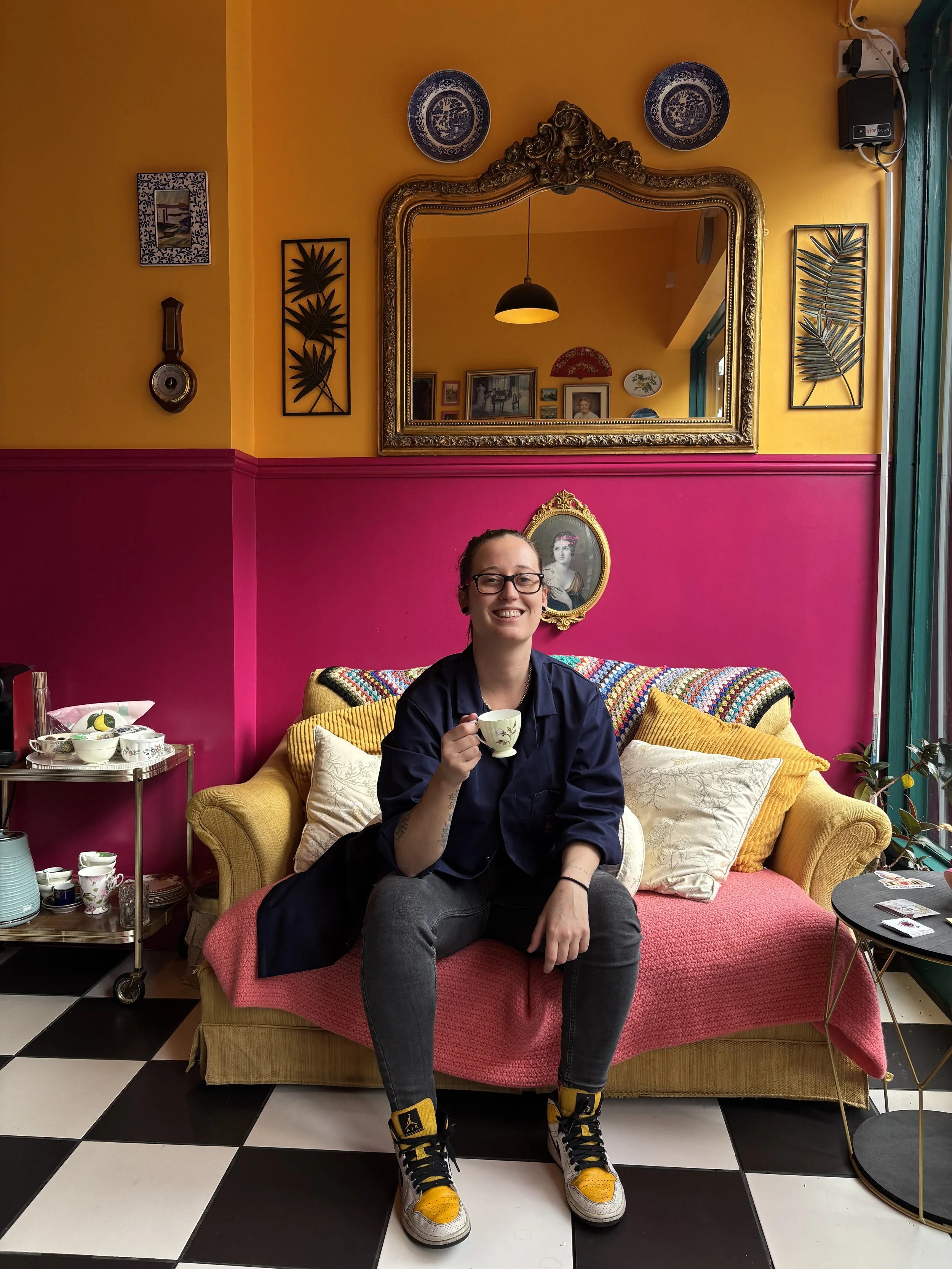 A young woman with glasses and a ponytail is sitting on a yellow sofa with a pink throw blanket, holding a teacup and smiling at the camera. The background features a colorful interior with a large ornate mirror, framed pictures, and decorative plates on a yellow and pink wall, with a checkered black and white floor.
