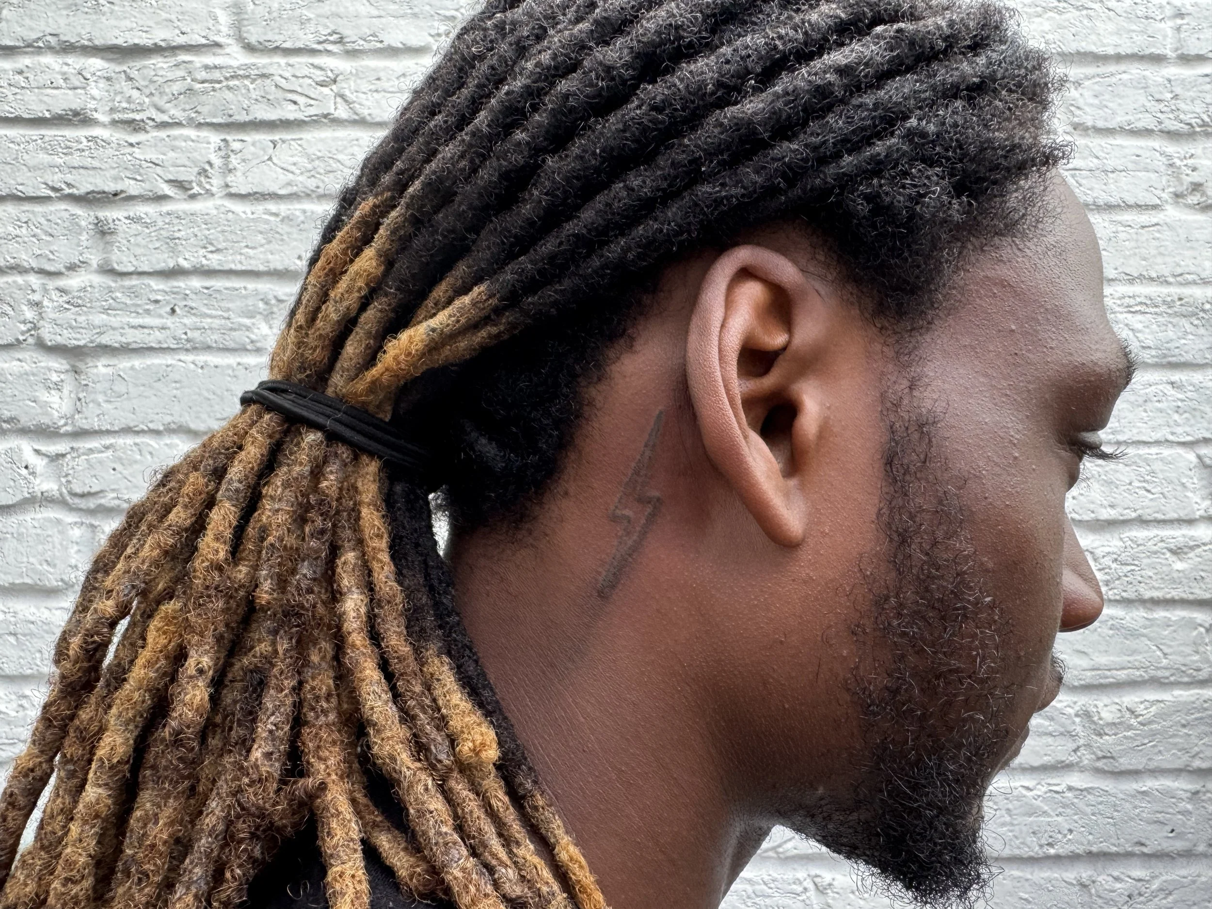 Close-up side profile of a person with dreadlocks tied back, a lightning bolt tattoo behind the ear, and a textured white brick wall in the background.