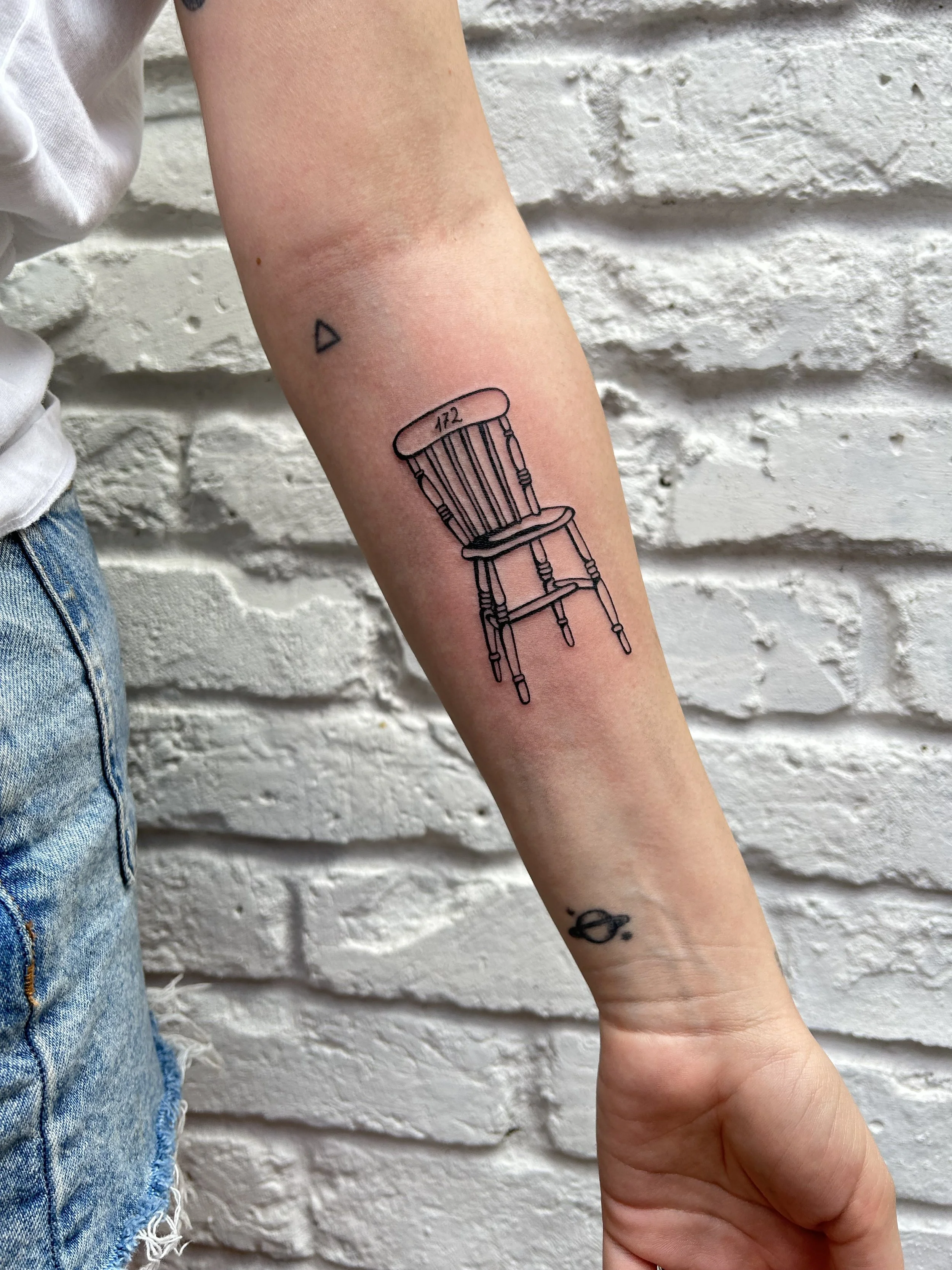 A tattoo of a simple, outline drawing of a wooden chair located on the inner forearm of a person. The chair has the number 472 written on the backrest, and there are small accompanying tattoos of a triangle above and a planet with rings below.