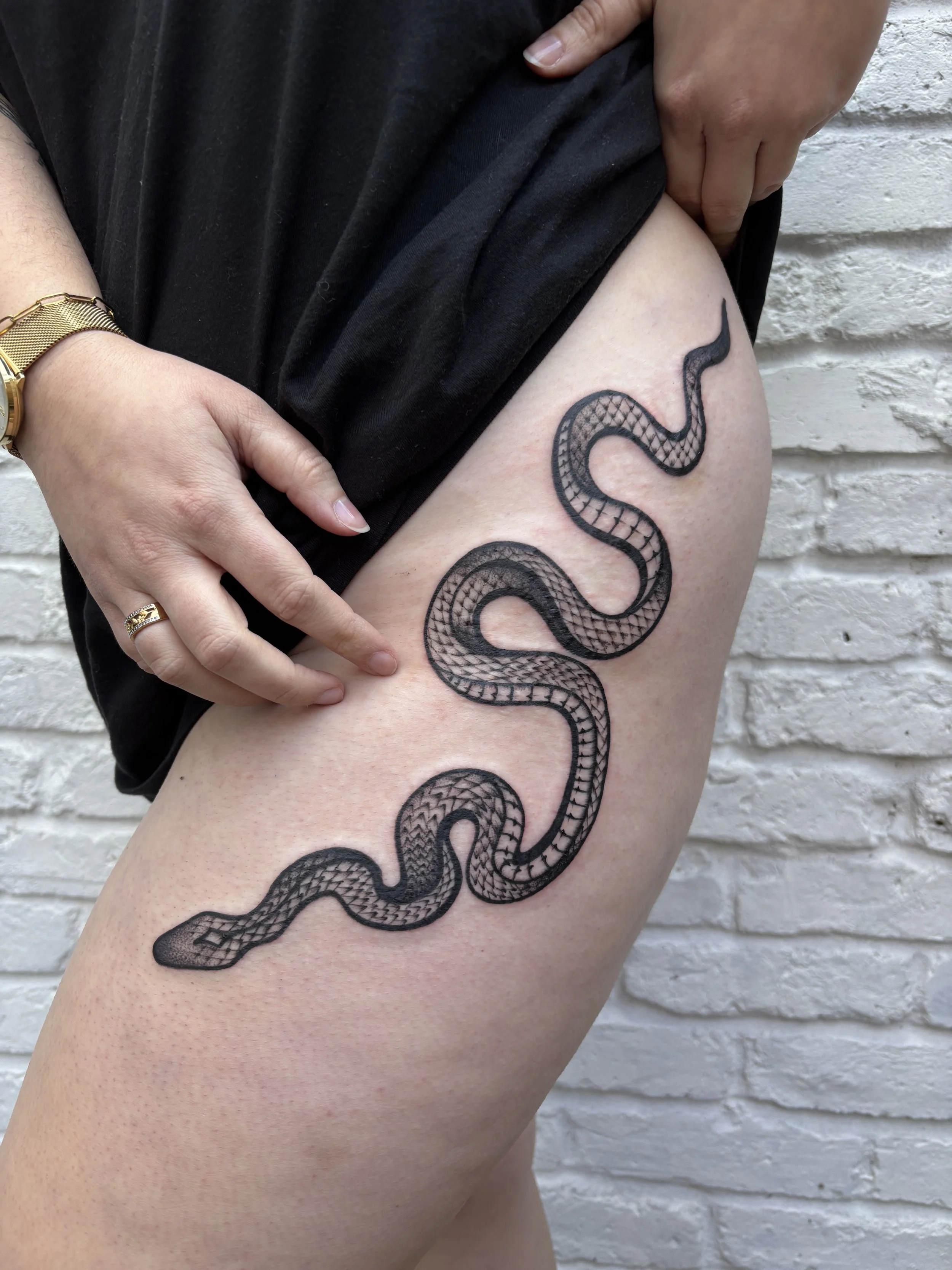 A person with a snake tattoo on their thigh, pointing at it while wearing a gold watch and ring, standing against a white brick wall.