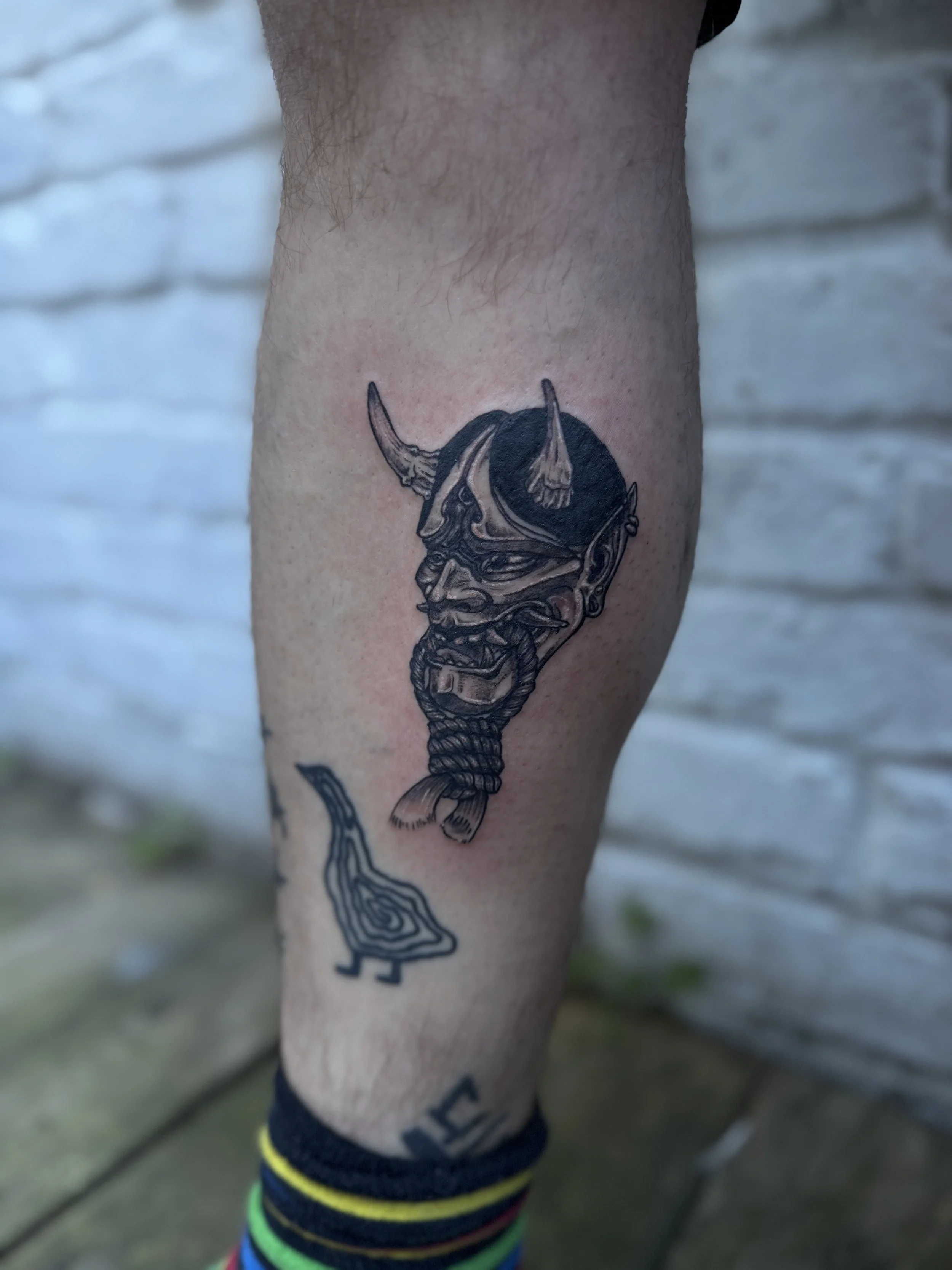 Tattoo of an oni mask with horns and a fierce expression, with a small abstract design below on a person's calf.