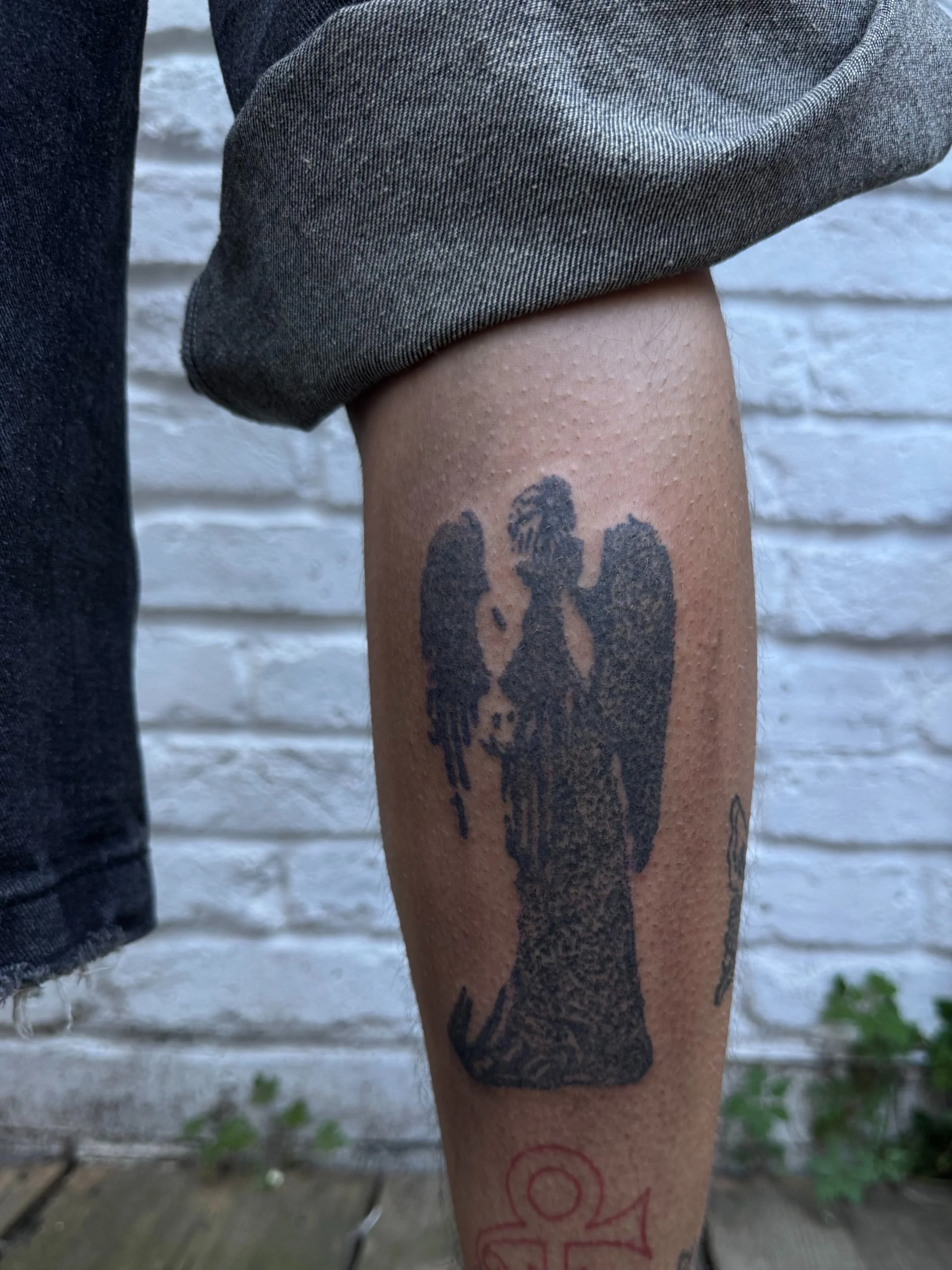 A tattoo of an angel with large wings, standing with its head bowed, on a person's calf. The person is wearing gray rolled-up pants and black shorts, with a white brick wall in the background.