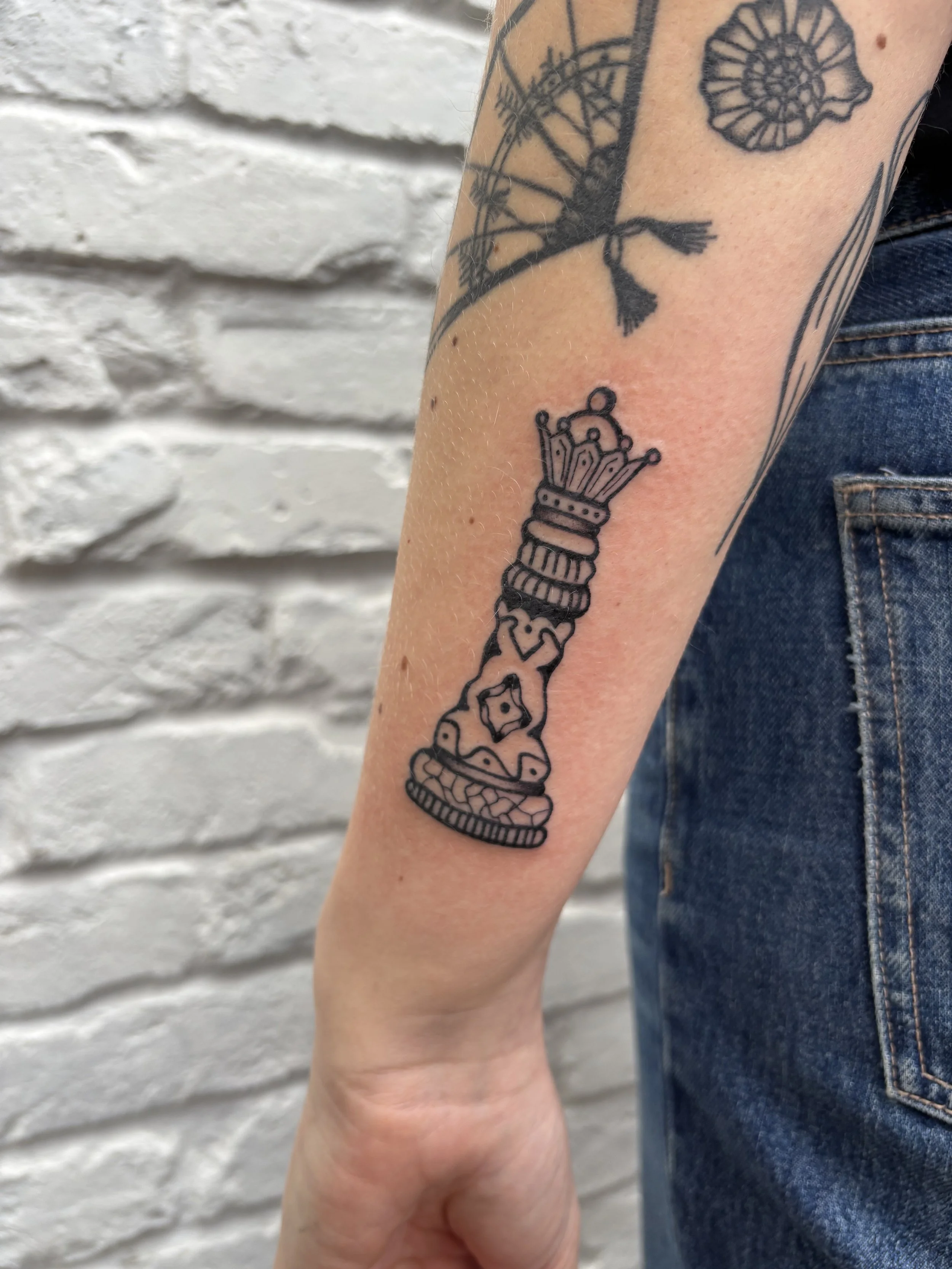 Tattoo of a stylized chess king piece on the forearm, with other tattoos visible in the background, against a white brick wall.