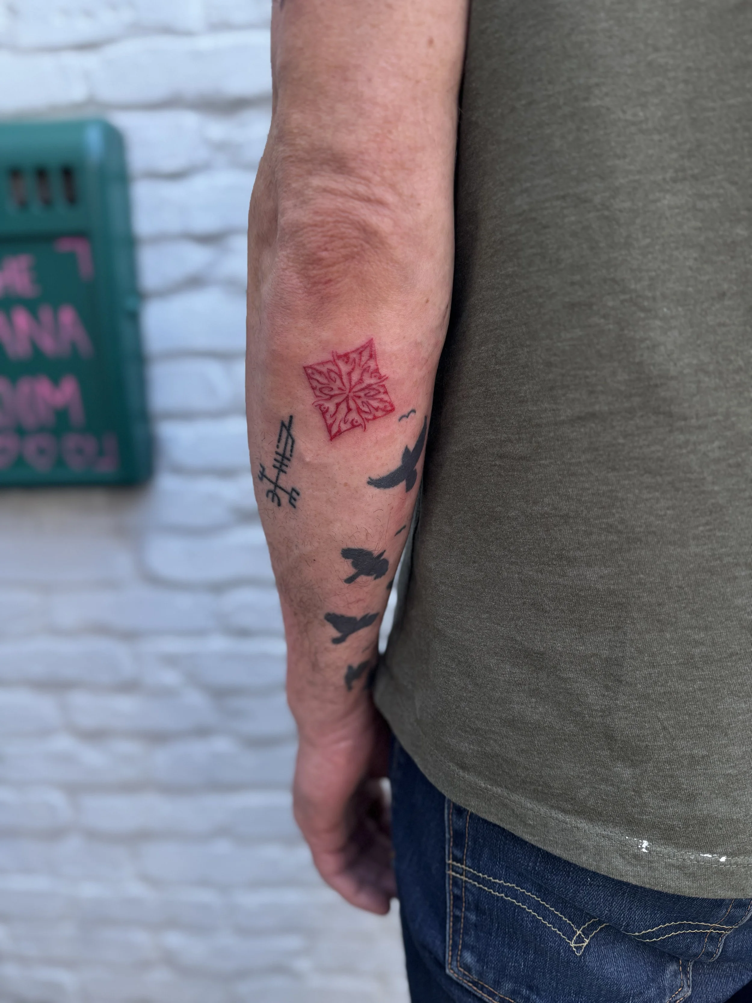 A person's arm with multiple tattoos, including a red geometric design, black birds, and small symbols, against a white brick wall background.