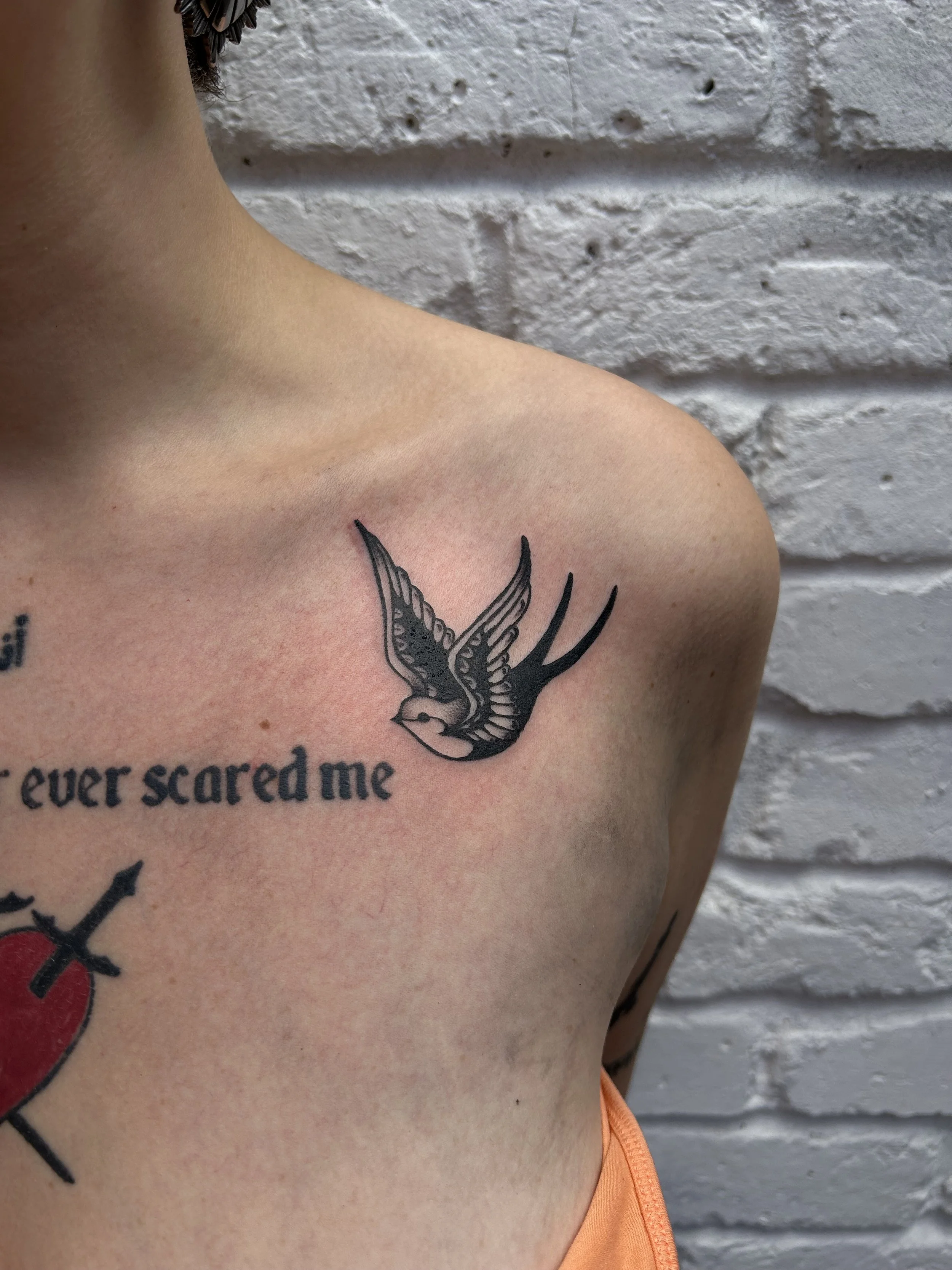 Tattoo of a flying swallow bird on upper chest near shoulder, with partial text that says 'ever scared me' visible underneath.