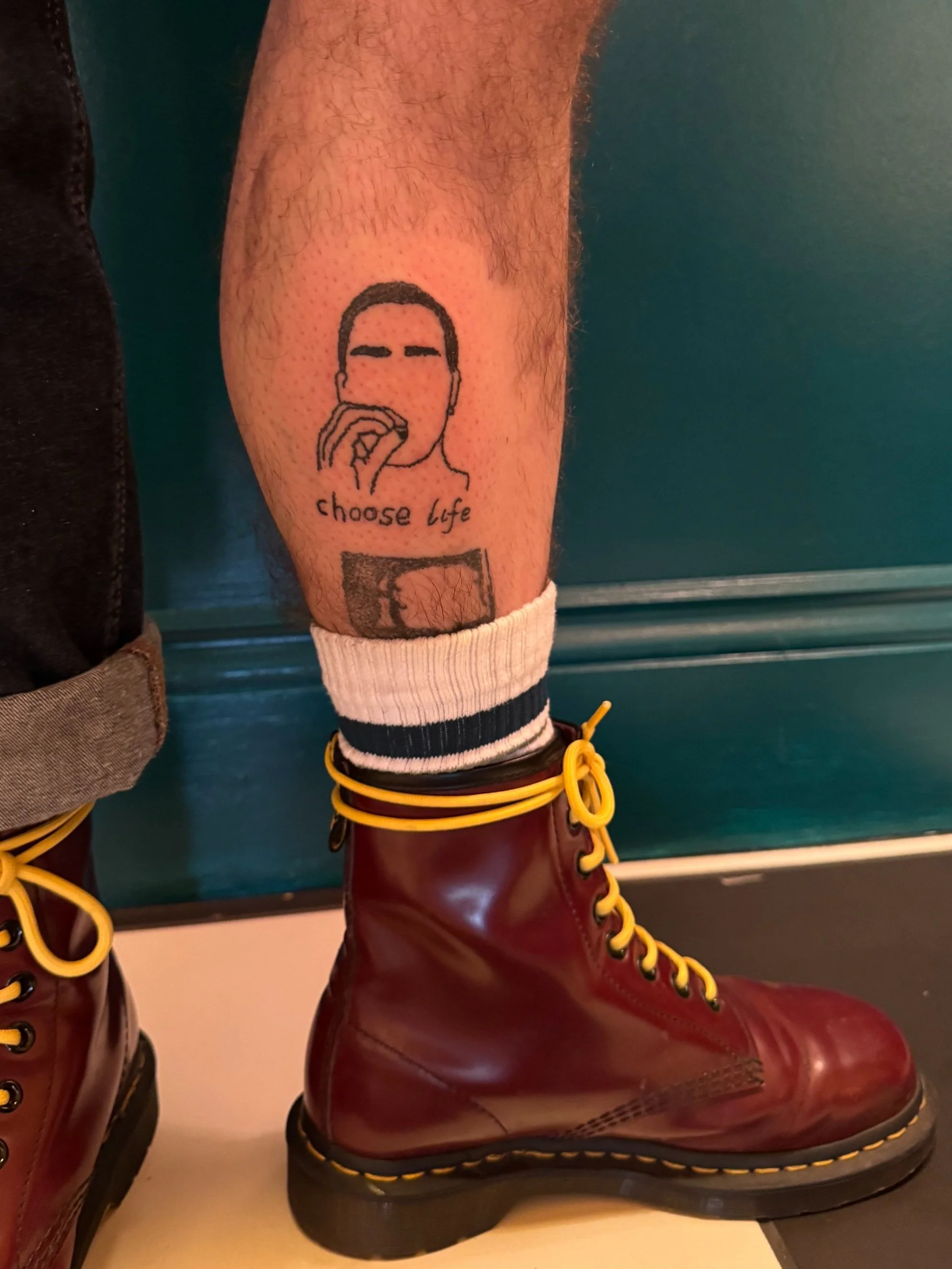 Tattoo of a person's face with blank eyes and nose, holding a hand to their mouth, with the words "choose life" underneath, on a person's lower leg near a brown boot and striped sock.