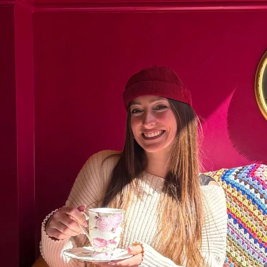 A woman smiling and holding a floral teacup with a saucer in a cozy room with a pink wall and a colorful blanket.
