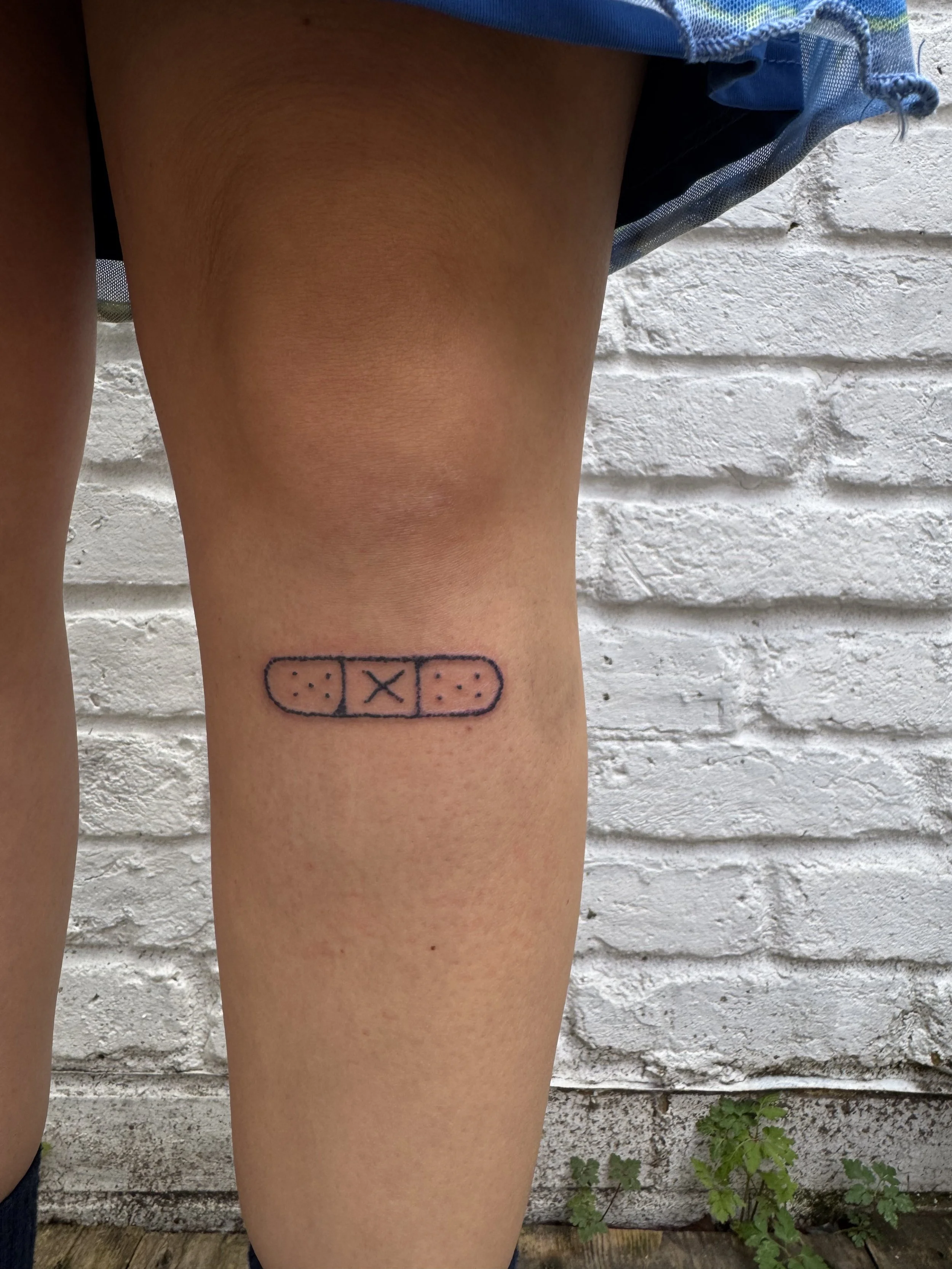 Tattoo of a pill capsule with a band-aid pattern on a person's thigh, against a white brick wall background.