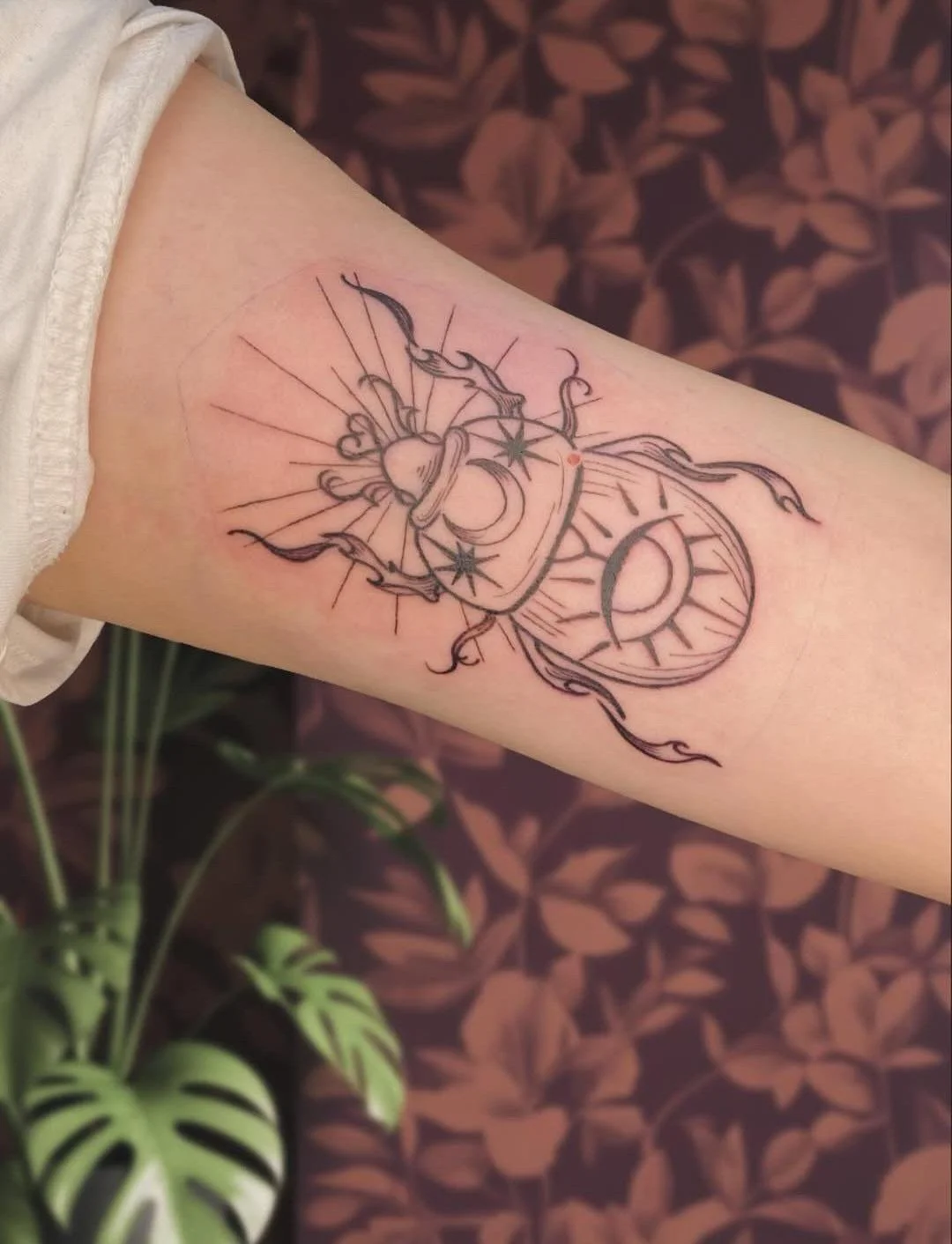 Line art tattoo featuring a potion bottle with a heart inside, surrounded by flames, stars, sun, moon, and radiating lines, on an inner forearm.