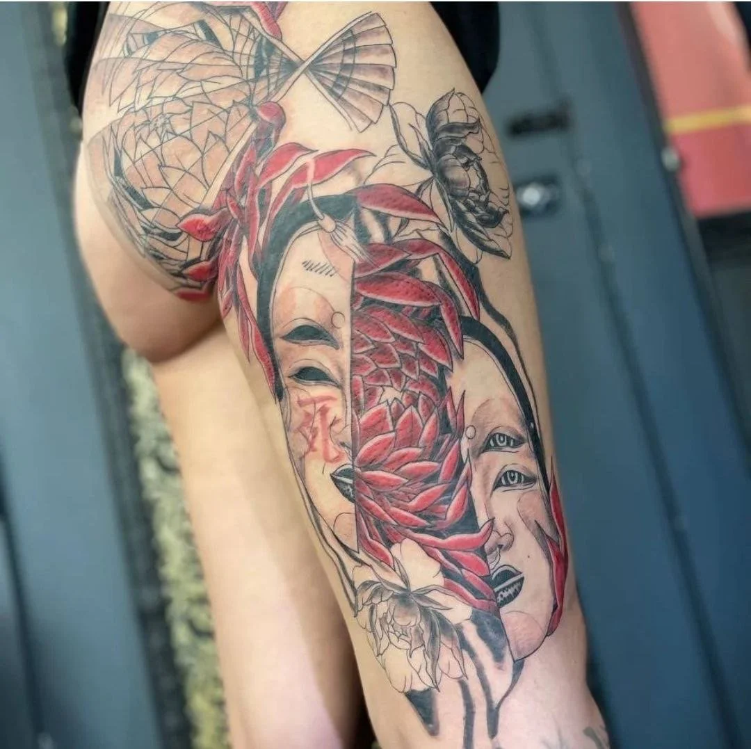 Tattoo of a woman's face with red leaves, flowers, a bee, and a fan, in a blend of black and red ink, on a person's arm and shoulder.