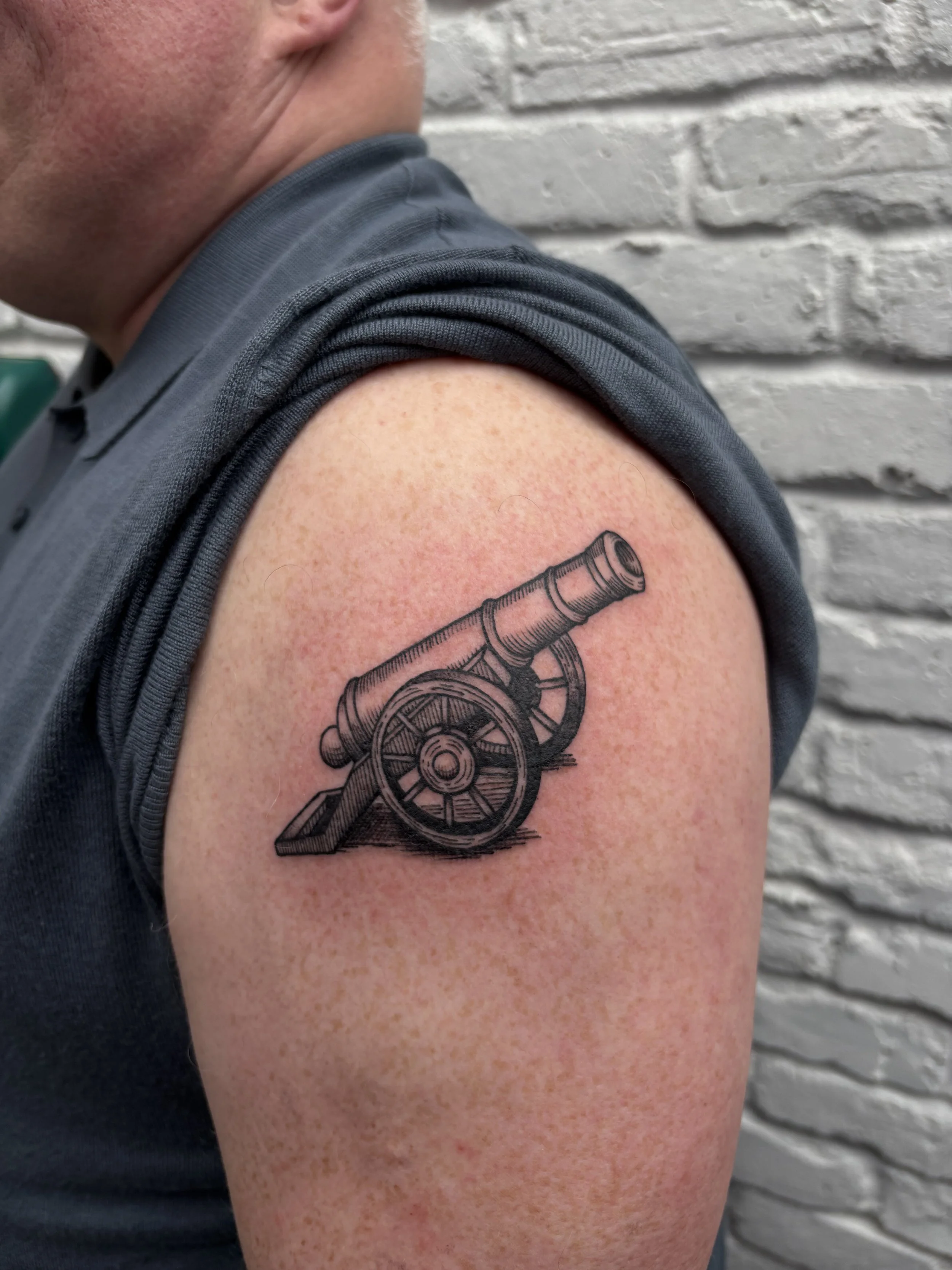 Tattoo of a black and gray cannon on a person's upper arm.