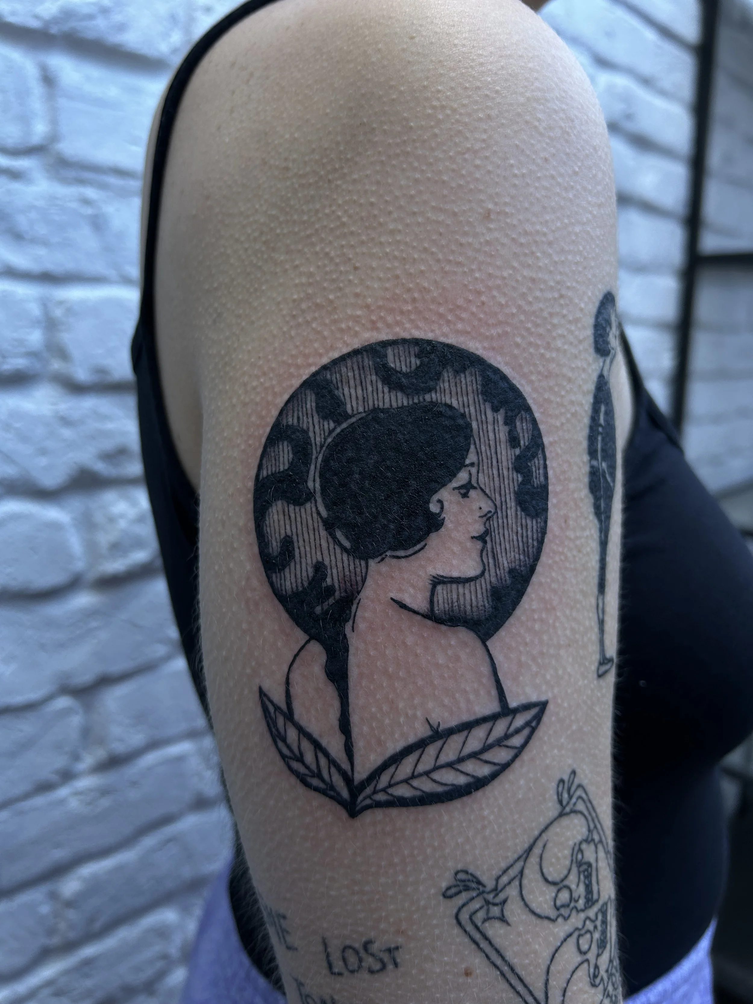 Tattoo of a woman's profile with dark hair, set within a circular background, with leaves at the base.