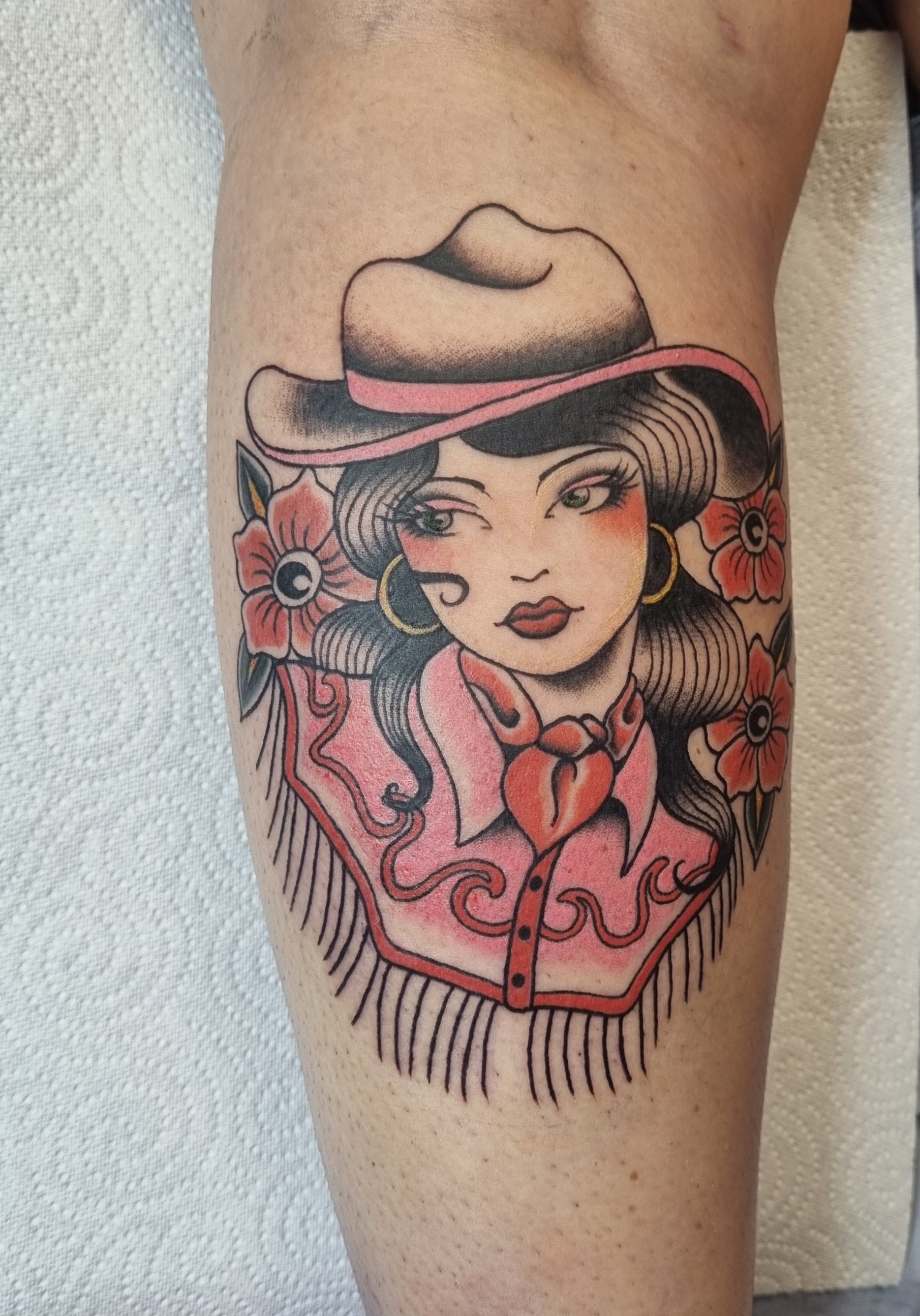 Tattoo of a woman with dark hair, green eyes, wearing a cowboy hat with pink band, gold hoop earrings, red lipstick, pink shirt with floral and Western-style design, and surrounded by red flowers with black centers.