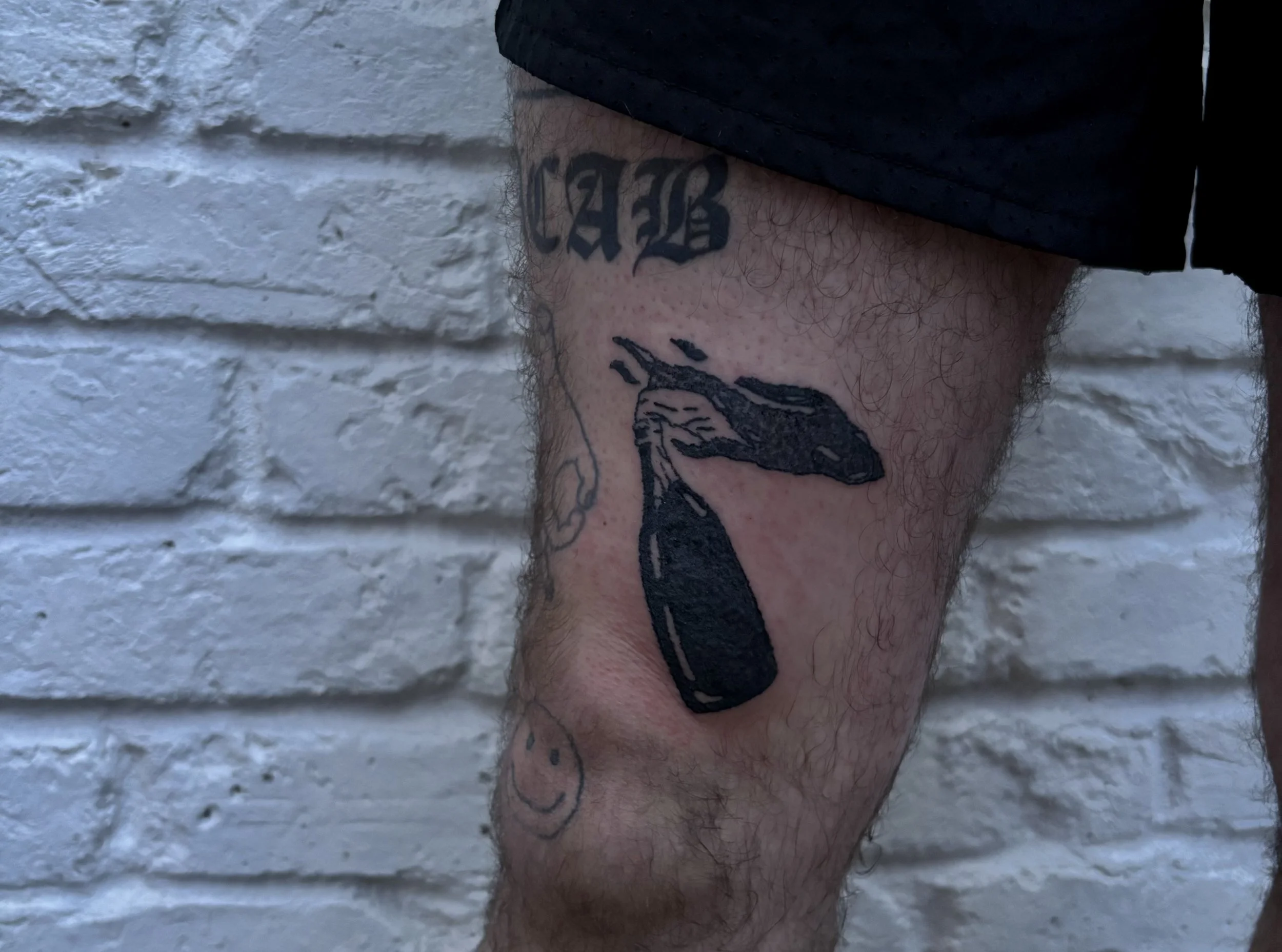 Tattoo of a cartoon-style bomb with a frowning animal face, on a person's thigh. The tattoo is black and the person has a tattoo above that says "CAB" in Gothic font.