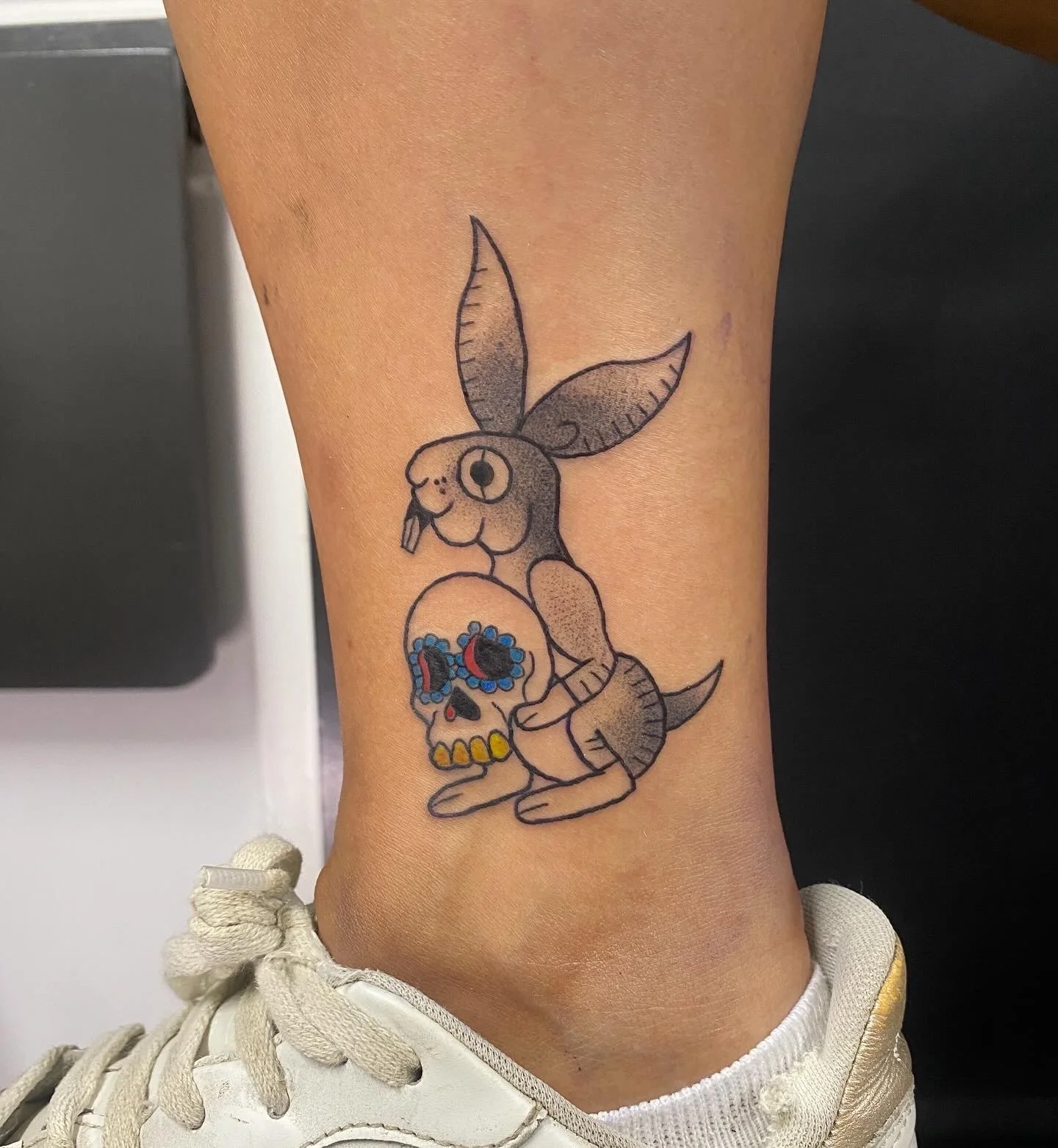 Tattoo of a bunny holding a decorated skull on ankle, with a white sneaker nearby.