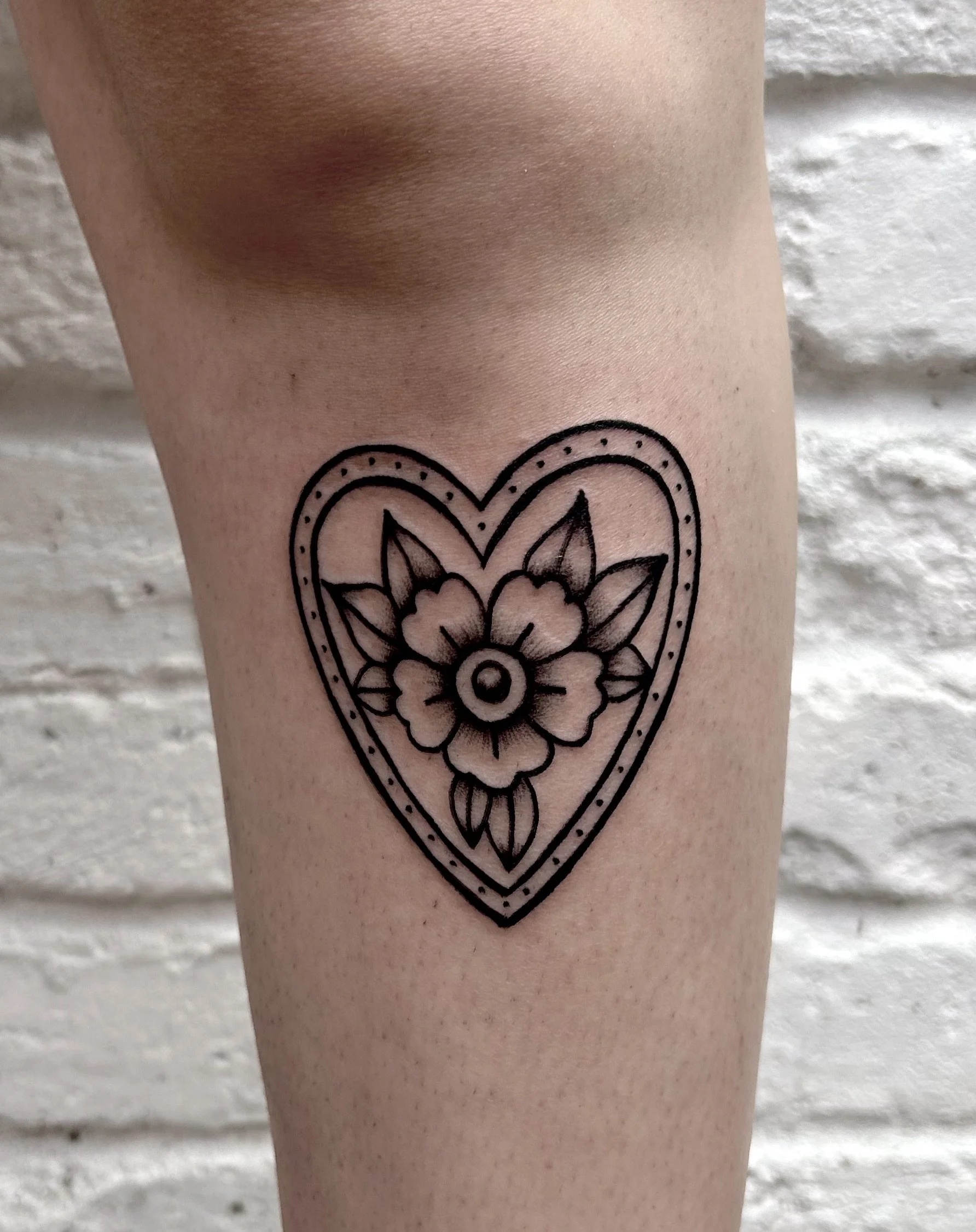 A black outline tattoo of a heart with a floral design inside, on a person's thigh against a white brick wall.