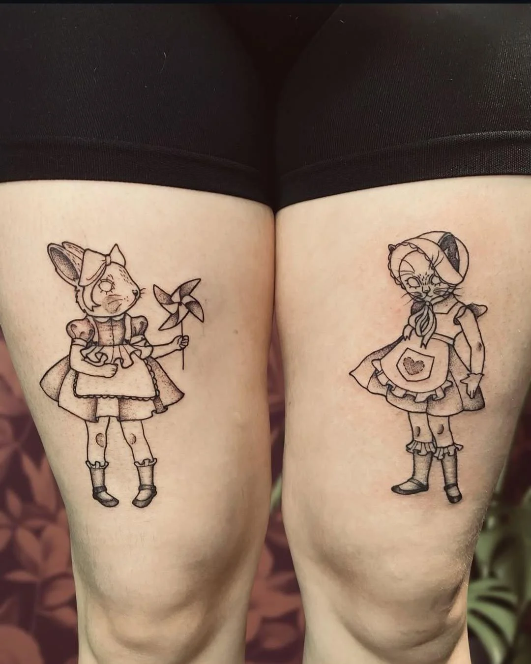 Tattooed thighs featuring black line art of anthropomorphic cats dressed as vintage or farmhouse characters, one holding a pinwheel on the left and the other wearing a bonnet and apron on the right.