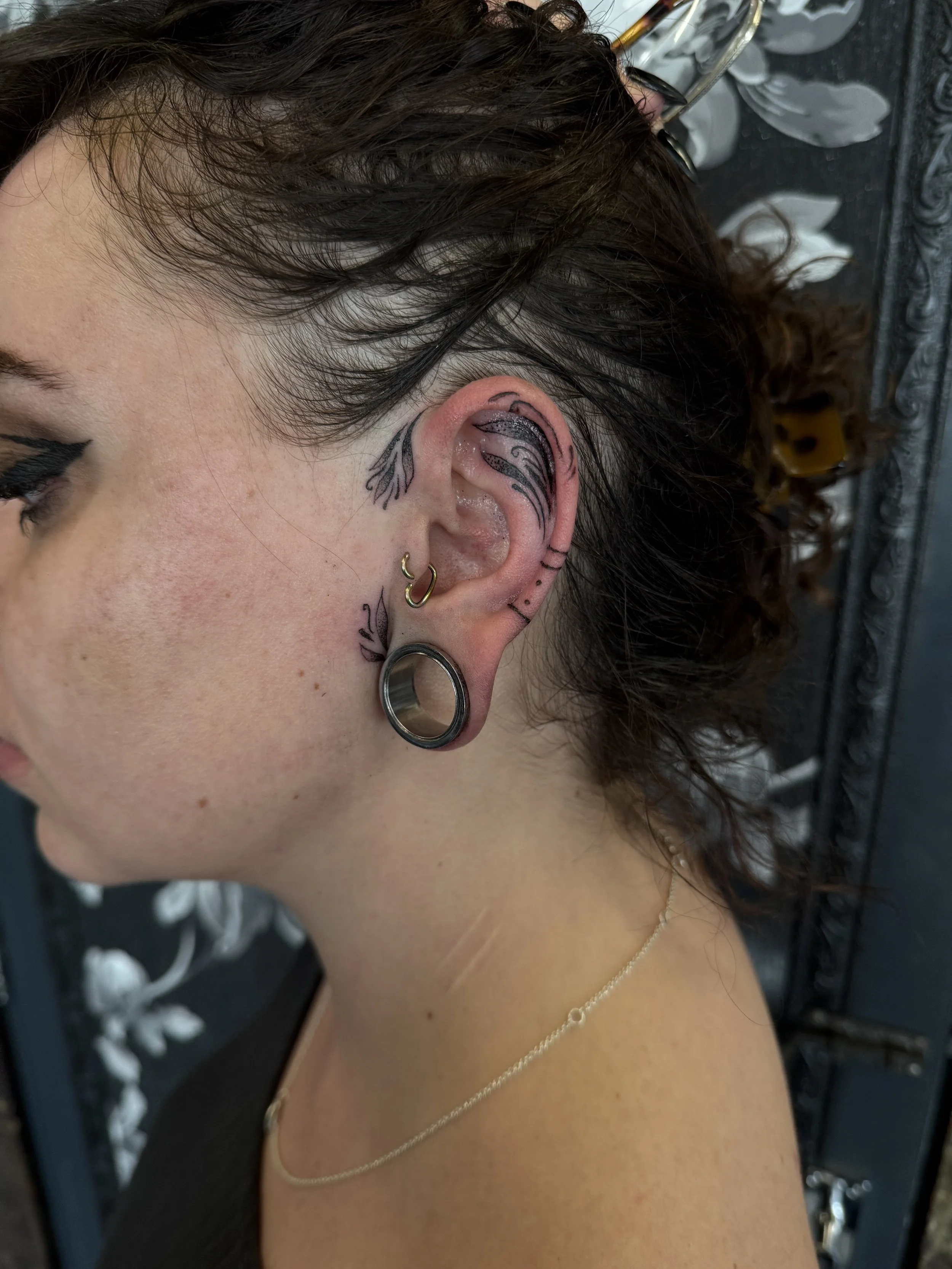 Close-up of a woman's head and shoulder, showcasing multiple ear piercings and tattoos behind the ear, hair tied in a bun, and a necklace on her shoulder.