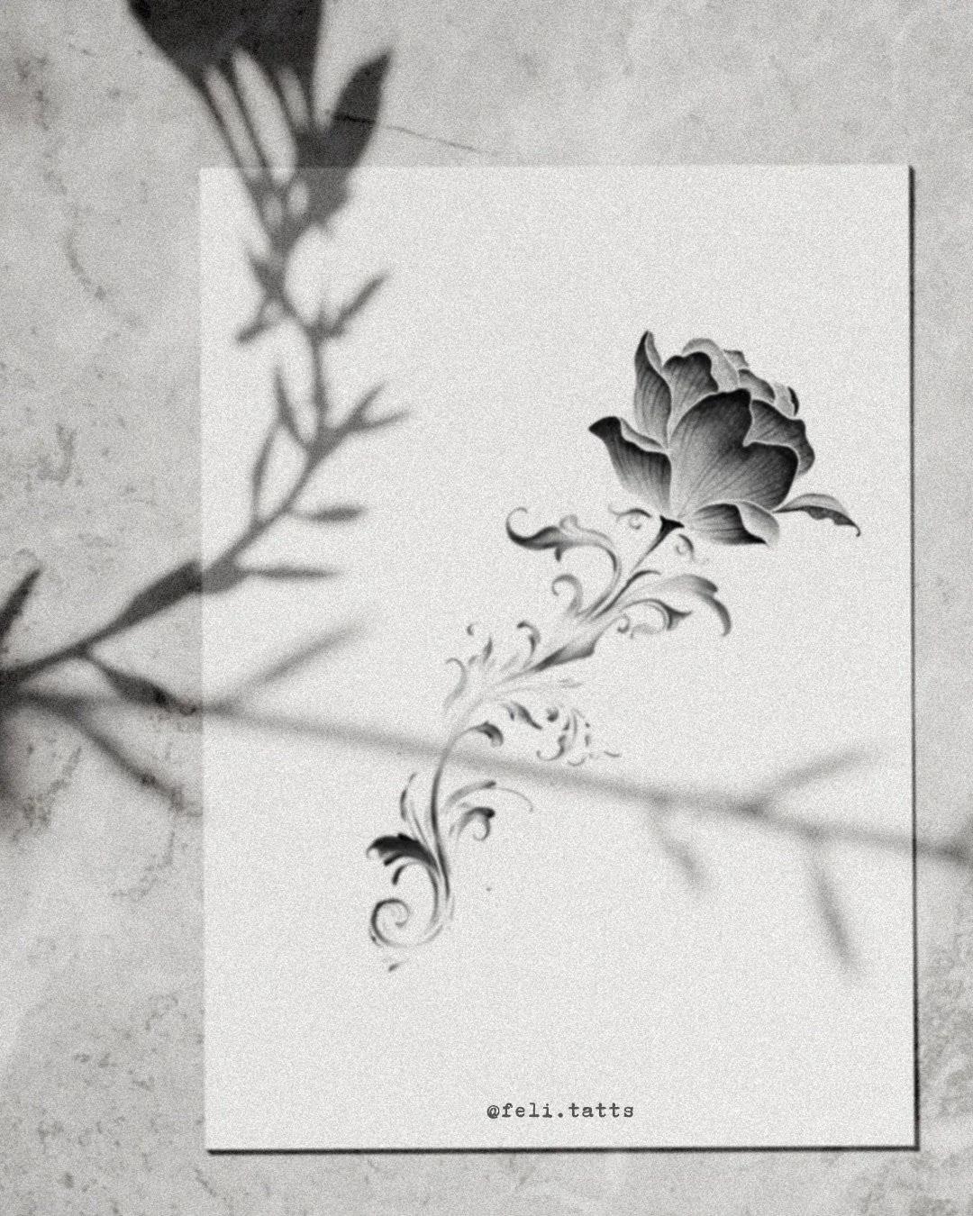 A grayscale tattoo design of a floral motif with a large flower and swirling leaves, on white paper with '@feli.tatts' written at the bottom.